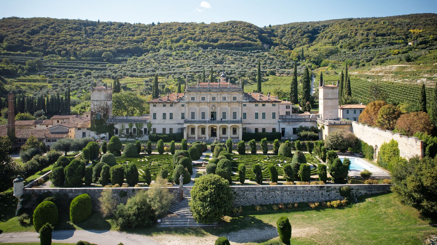Aerial view of a grand Italian villa with manicured gardens and cypress trees nestled in rolling Tuscan hills.