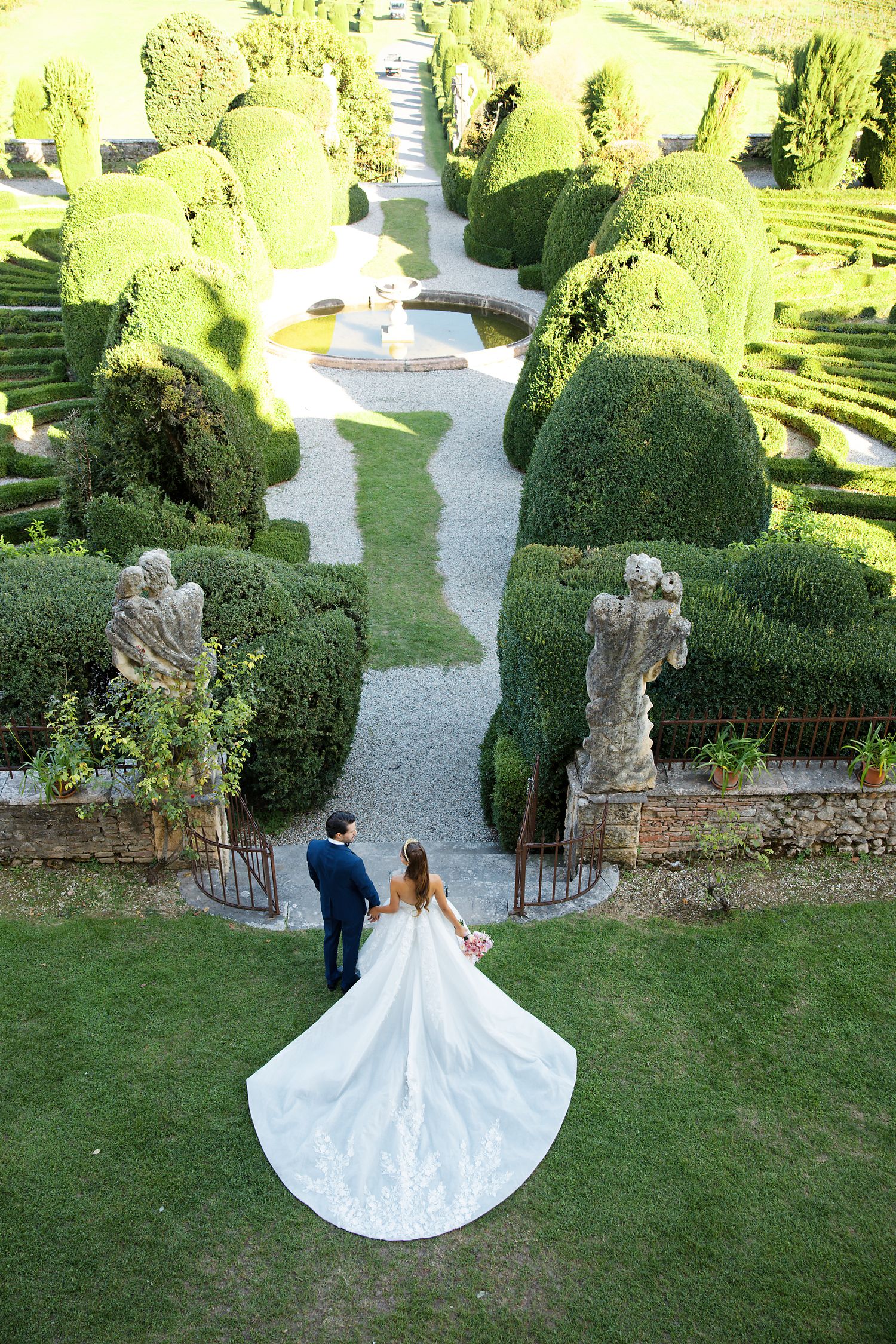 A couple stands at the entrance of an elegant formal garden with manicured topiary and winding pathways.