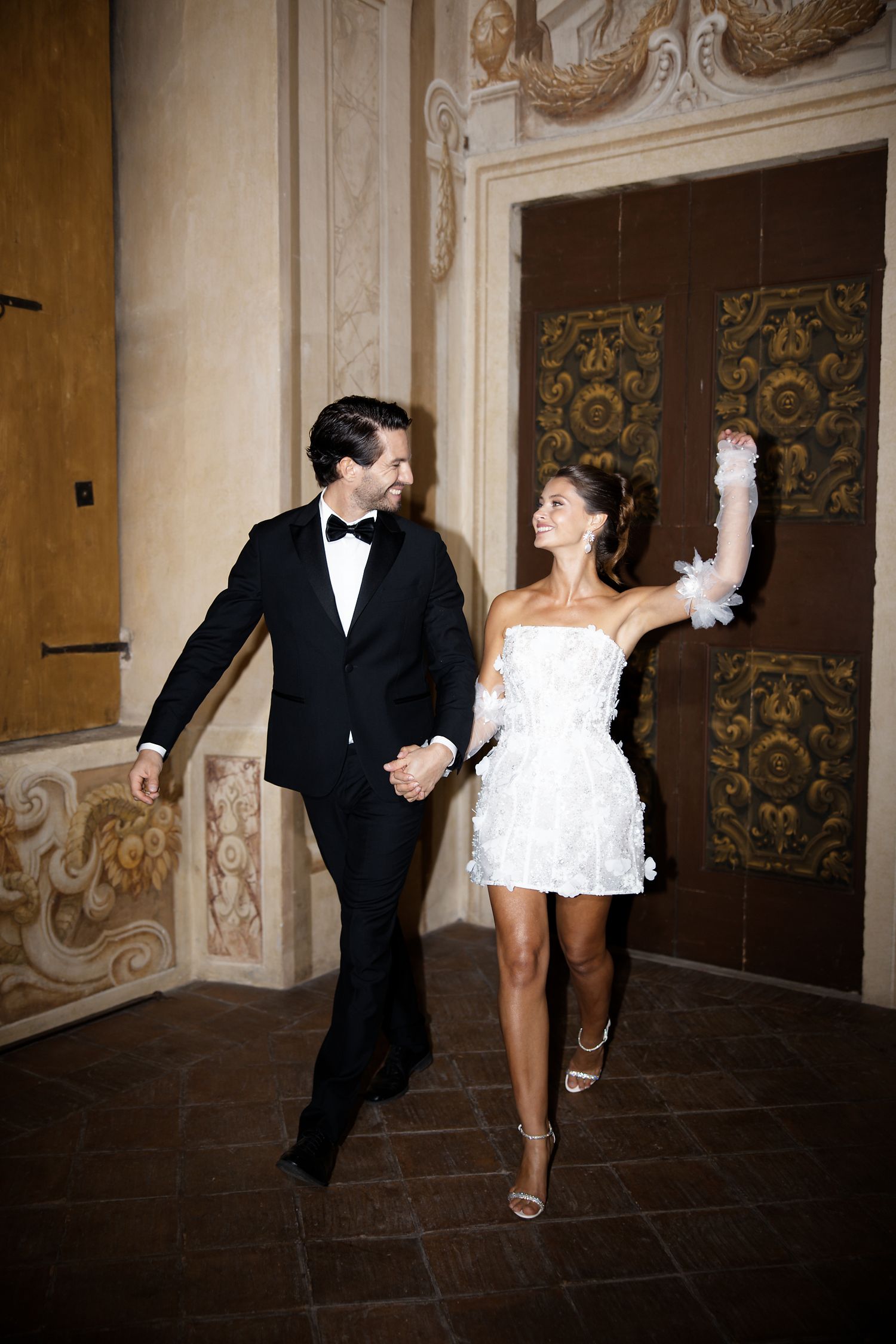 Dancing couple in formal attire moves through elegant hallway with ornate wall details.
