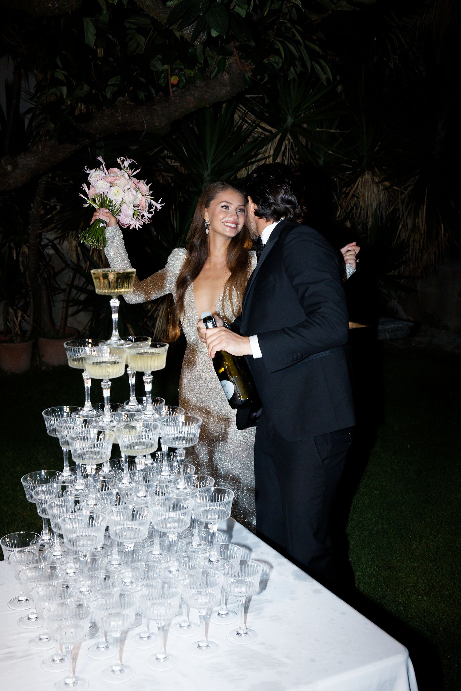Newlyweds share a moment by an elaborate champagne tower display with floral arrangements at their reception.