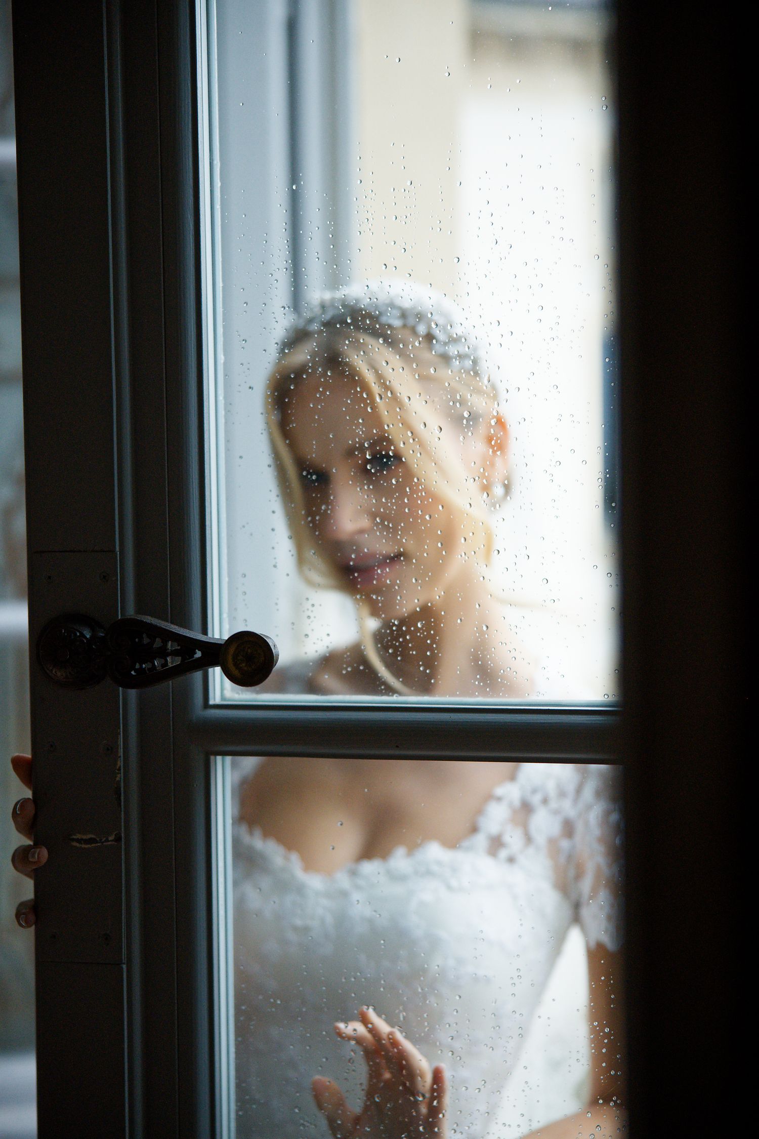 A dreamy portrait through a rain-speckled window of a bride in a delicate lace wedding dress.