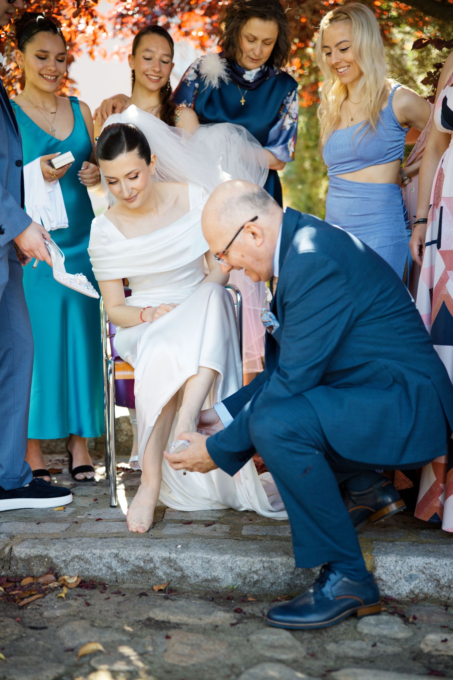 Wedding party gathered outdoors while someone adjusts the bride's shoe in autumn lighting.