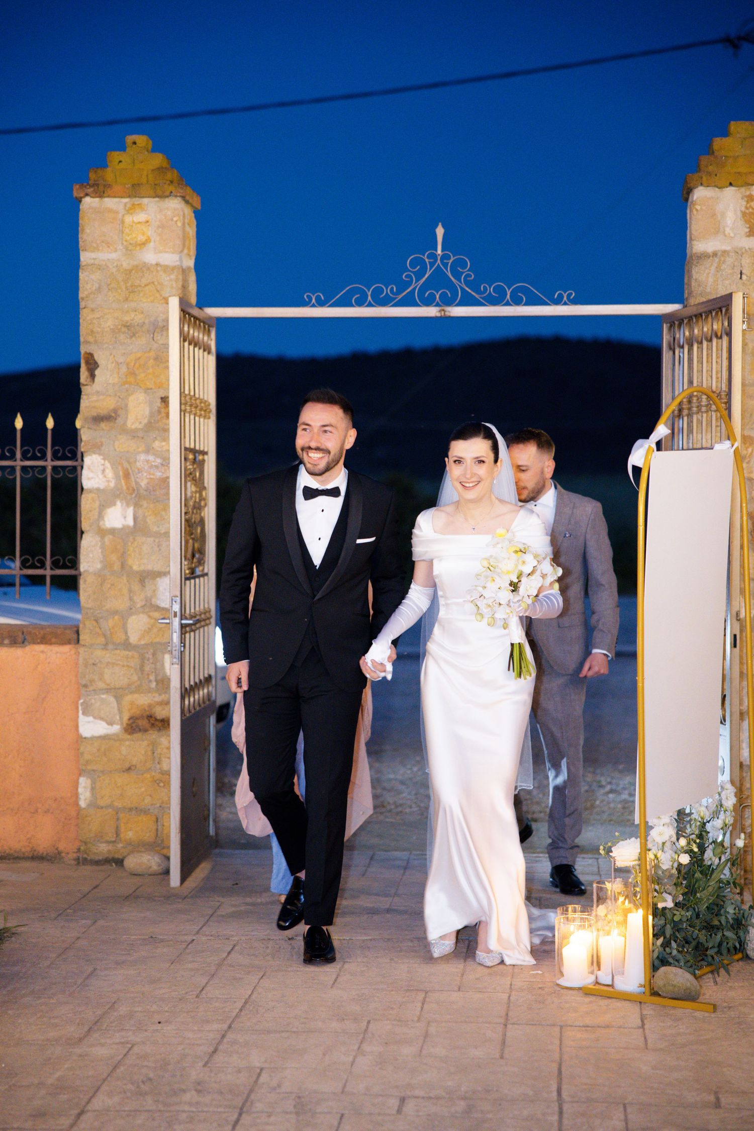 A wedding party enters through an ornate stone archway at dusk with lantern lighting along the entrance path.