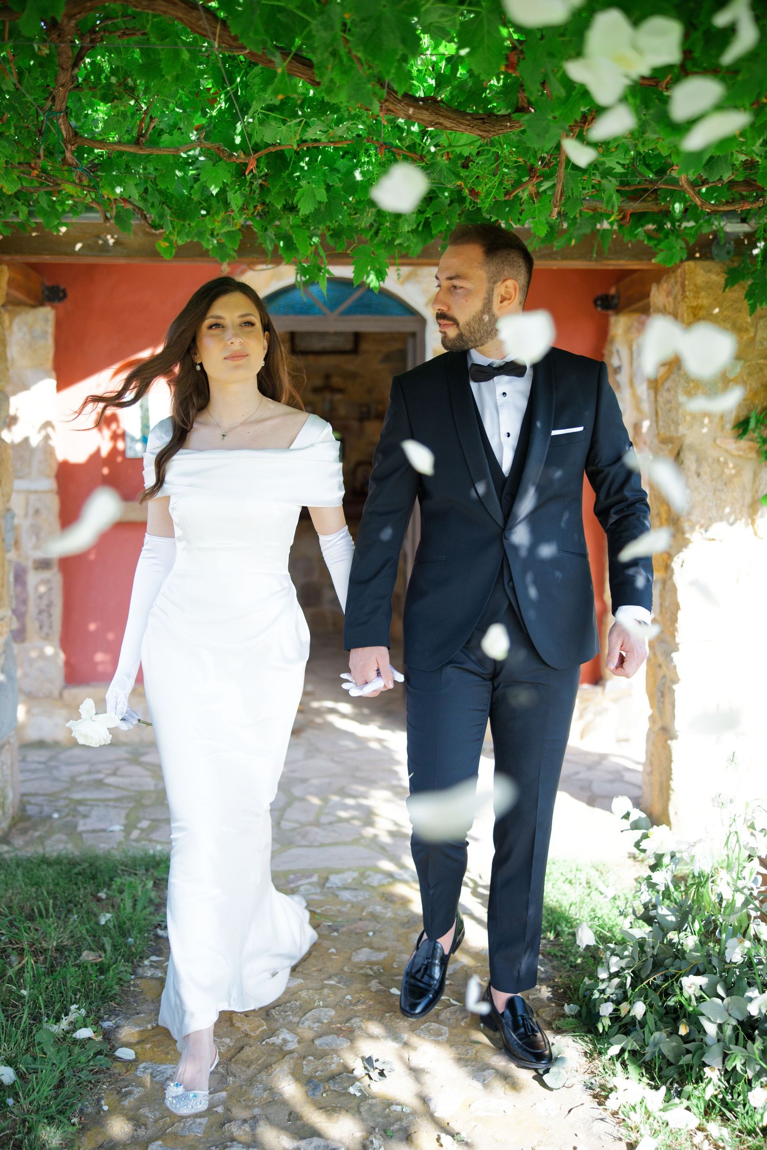 A romantic wedding couple holding hands walks under a floral archway in elegant white dress and navy tuxedo.
