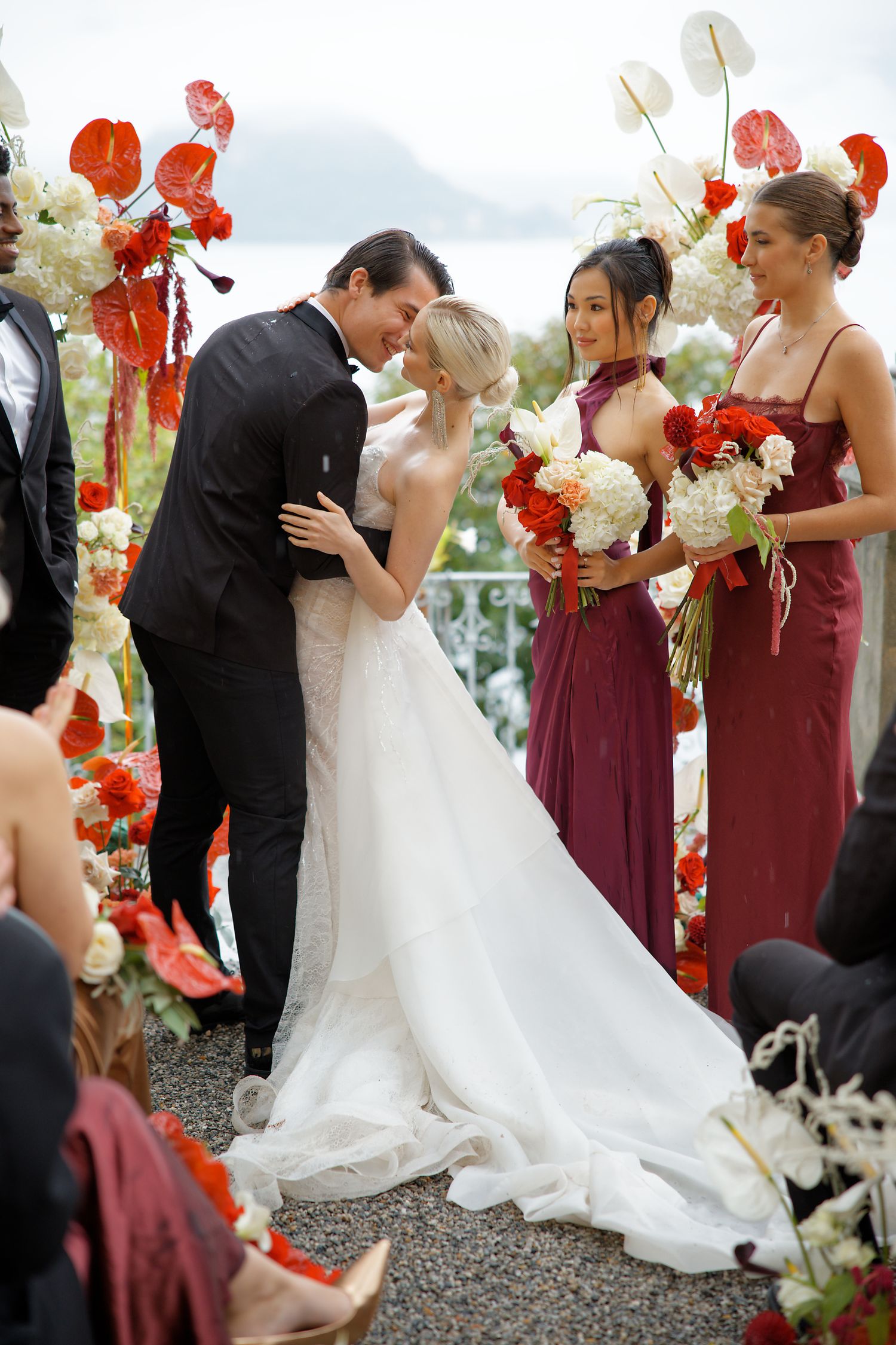 Wedding ceremony under a floral arch with red and white blooms and attendants in burgundy dresses.