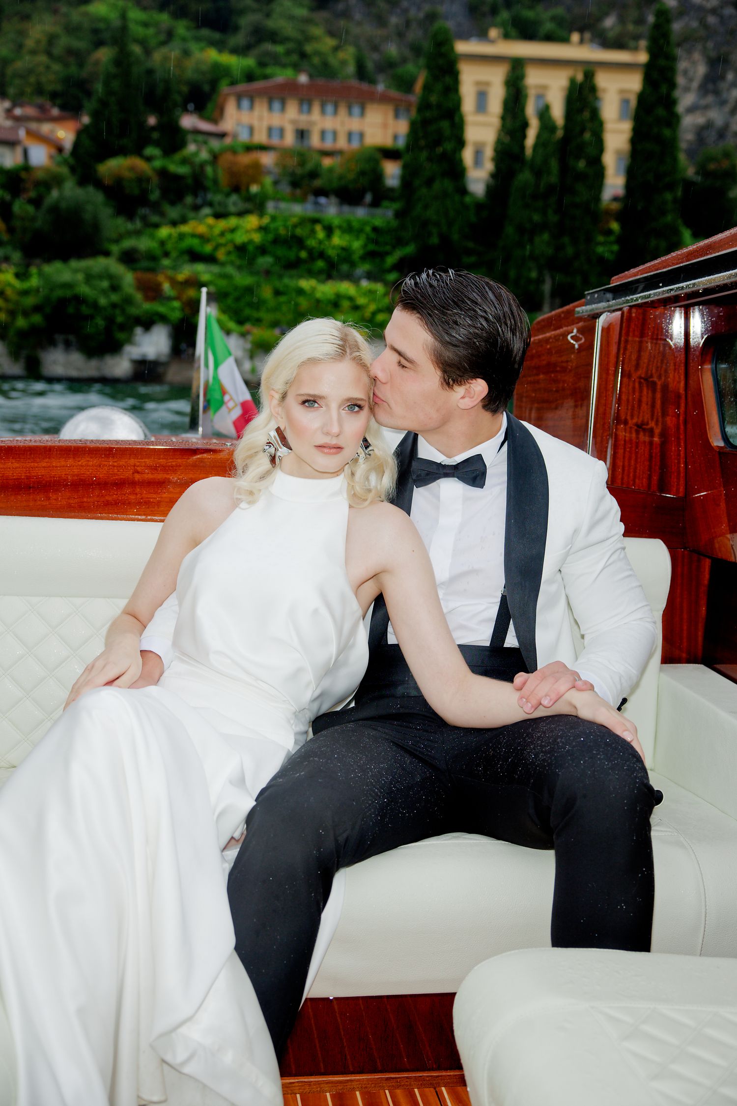 A romantic newlywed couple embraces on a classic wooden boat against a scenic Mediterranean coastal backdrop.