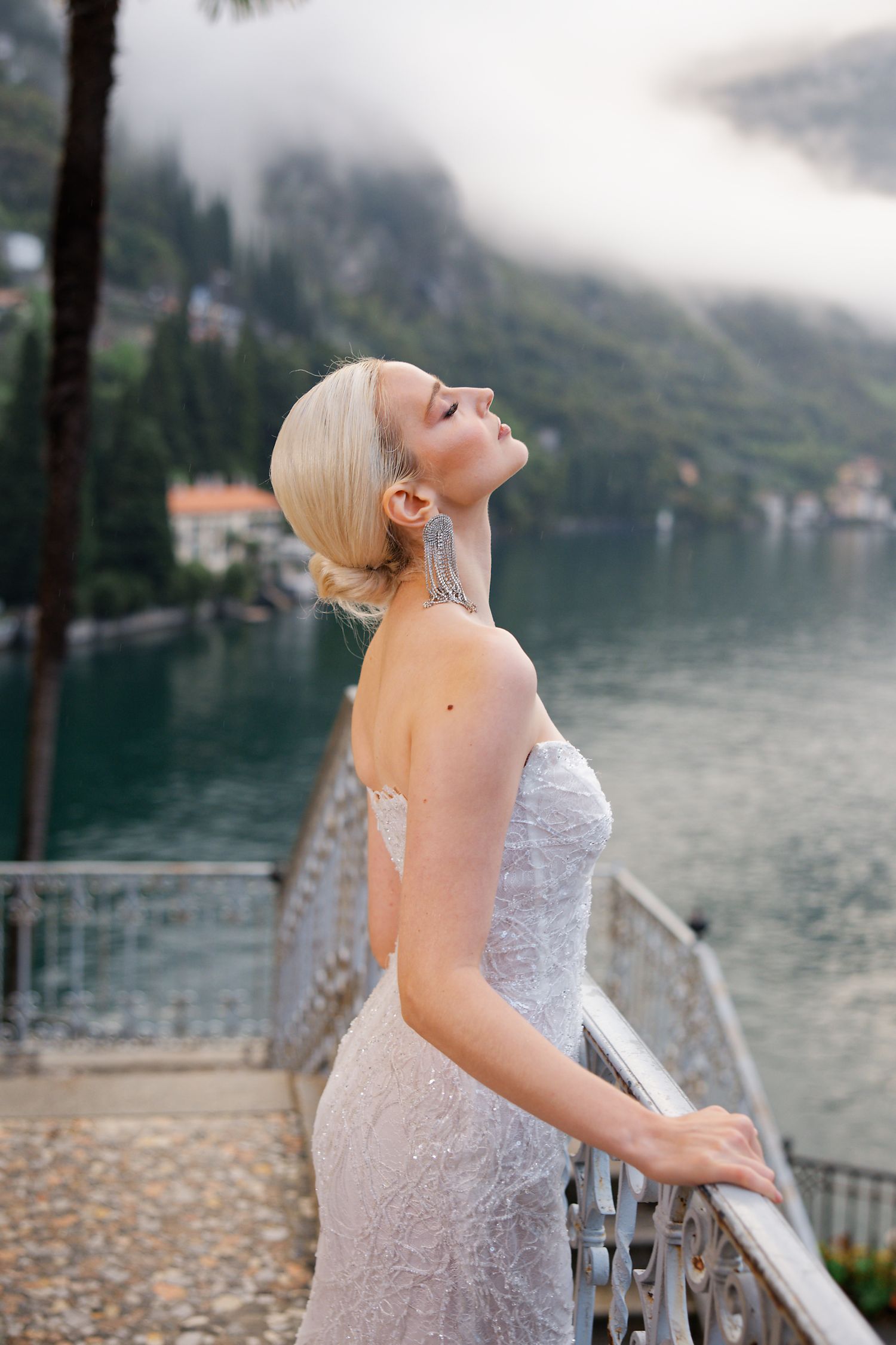A bride in a lace gown poses on a balcony overlooking a misty lake and mountains.