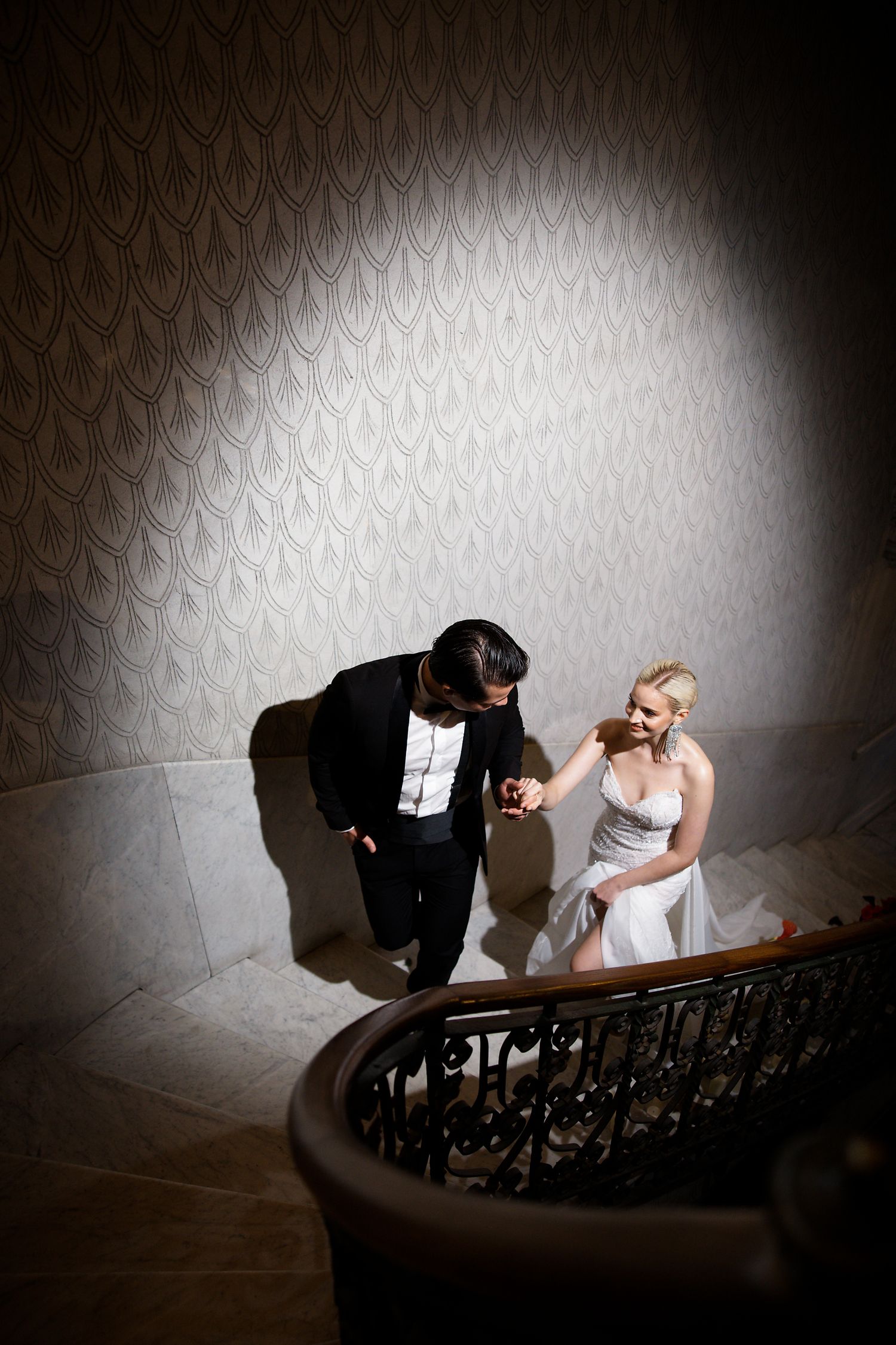 A couple in formal evening wear ascends an elegant staircase with ornate wallpaper and railing in dramatic lighting.