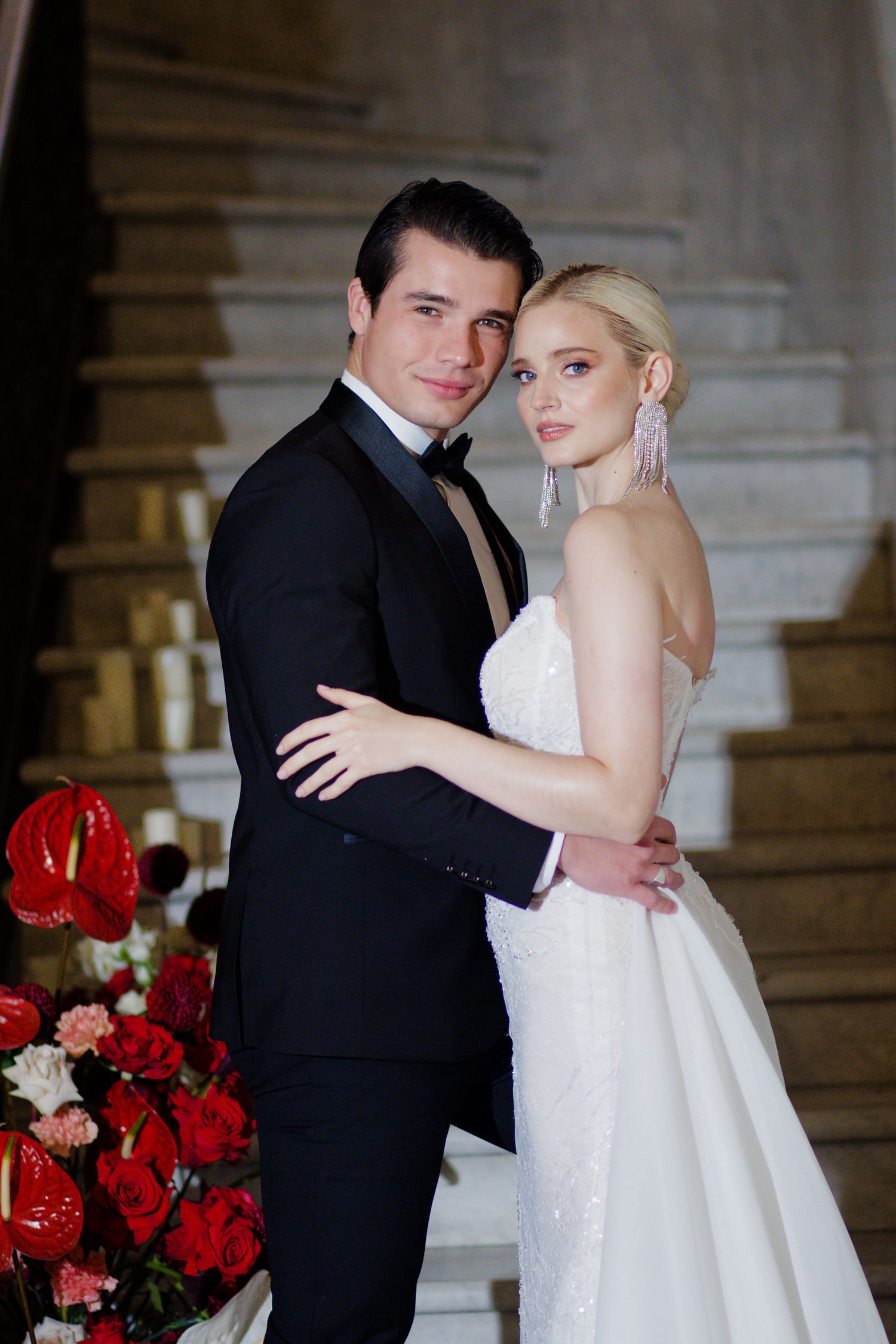 A couple poses elegantly on marble stairs with red floral arrangements in formal wedding attire.