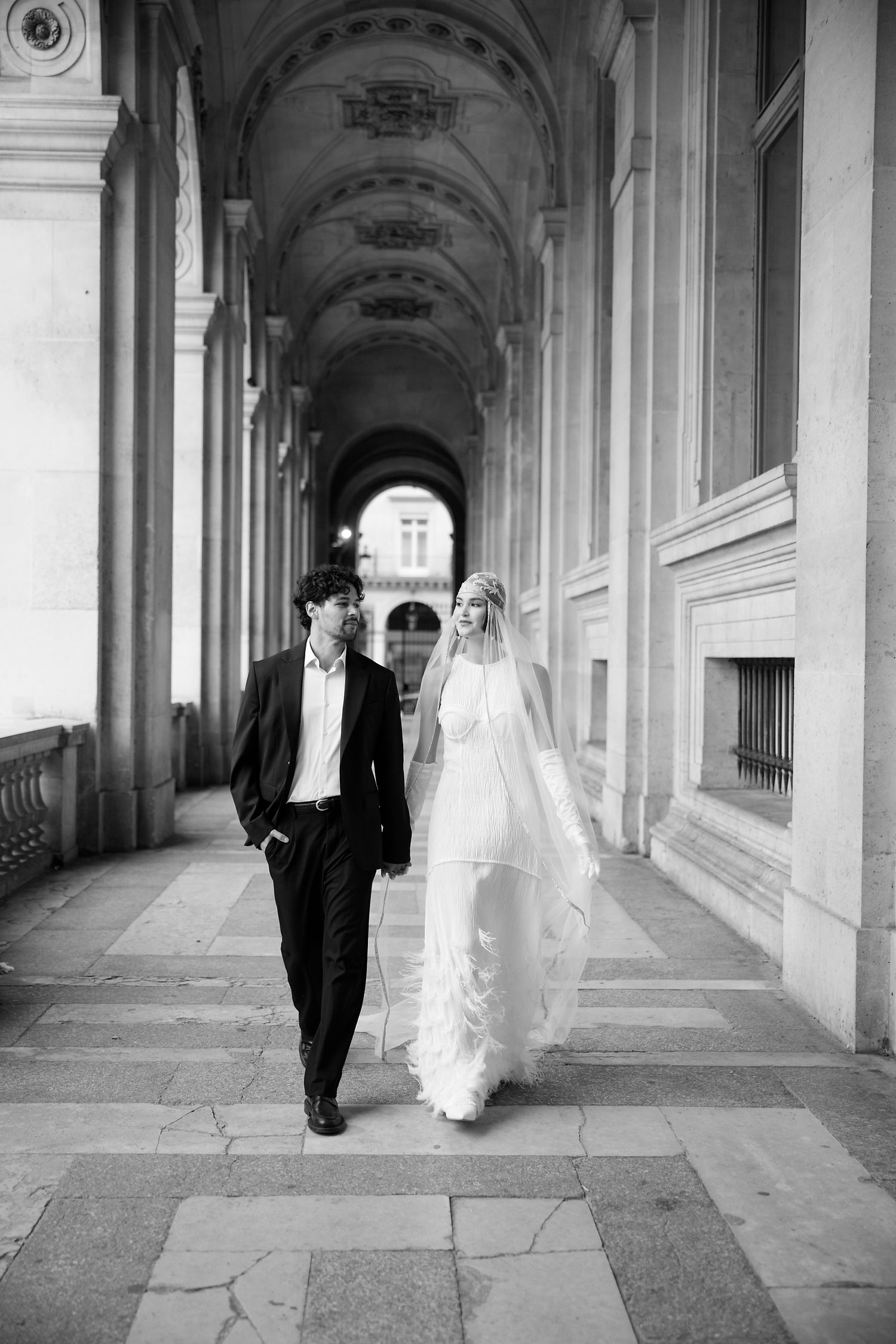 A bride and groom walk through an elegant arched corridor in black and white photography.