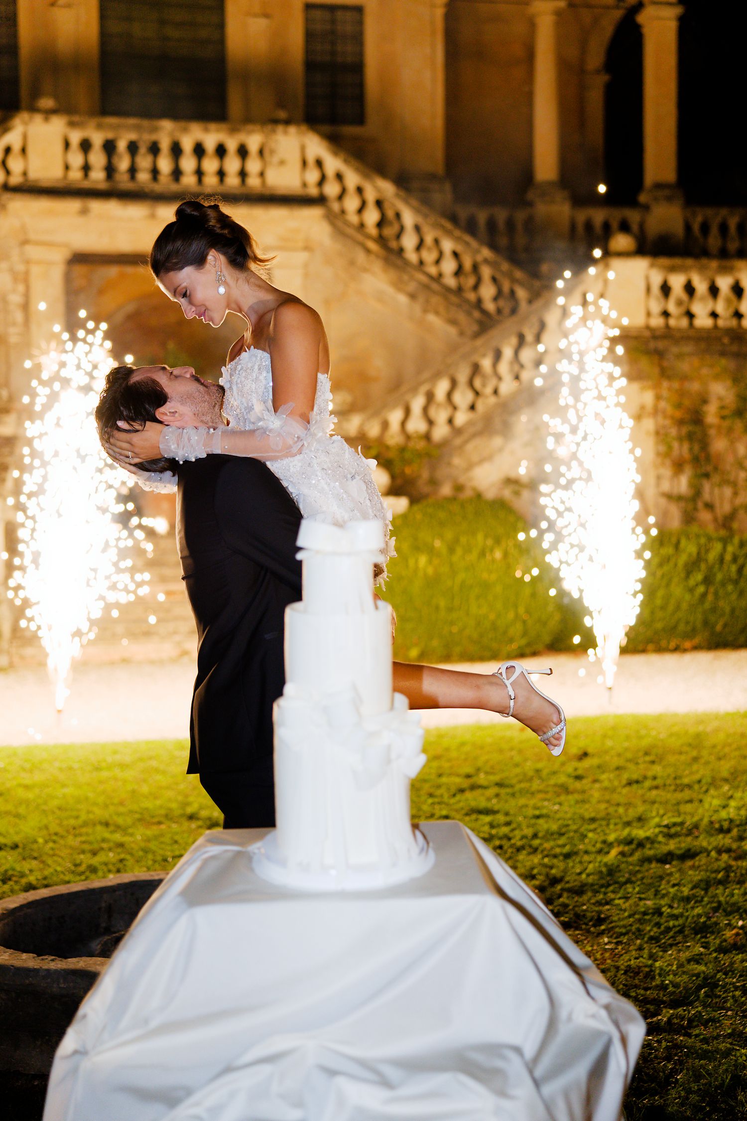 Romantic wedding cake cutting moment with sparklers at night in front of historic villa.