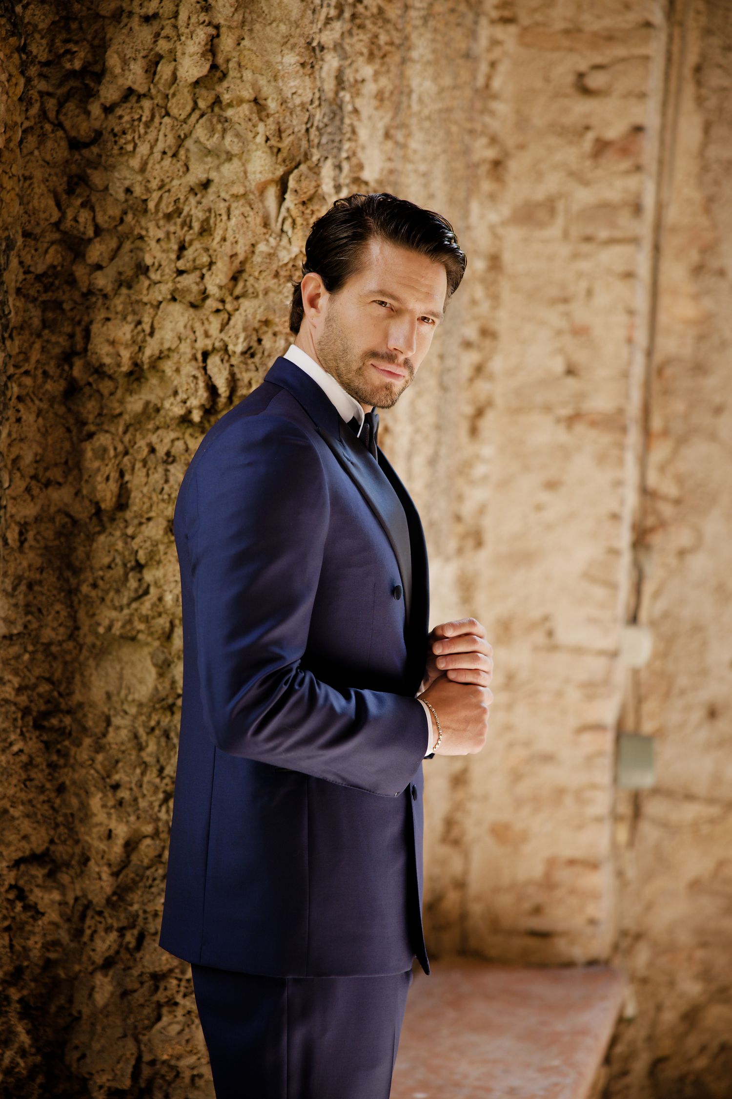 Elegant figure in a navy blue suit posed against a textured stone wall in warm lighting.