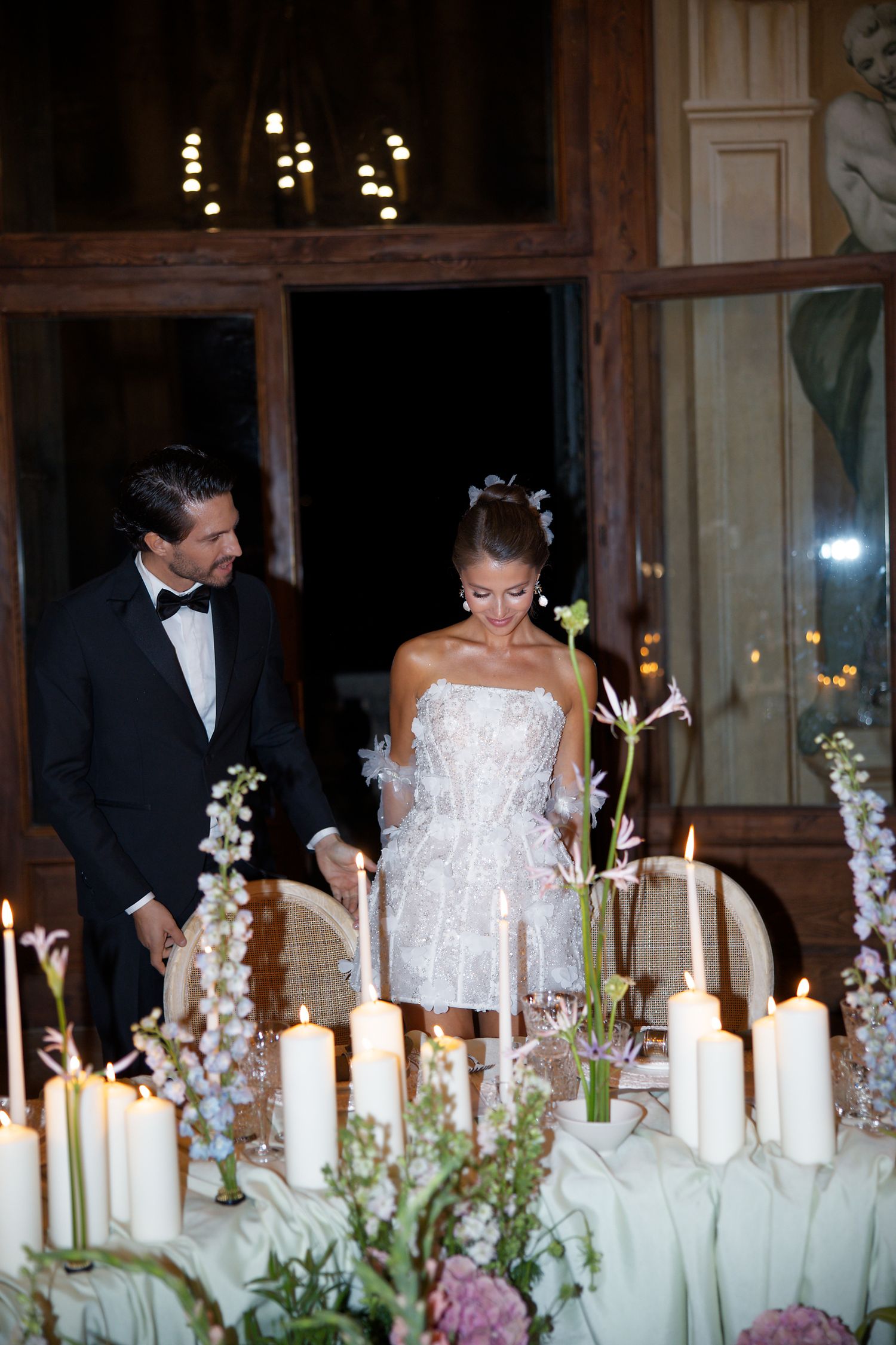 Couple stands by romantic candlelit table with white floral arrangements.