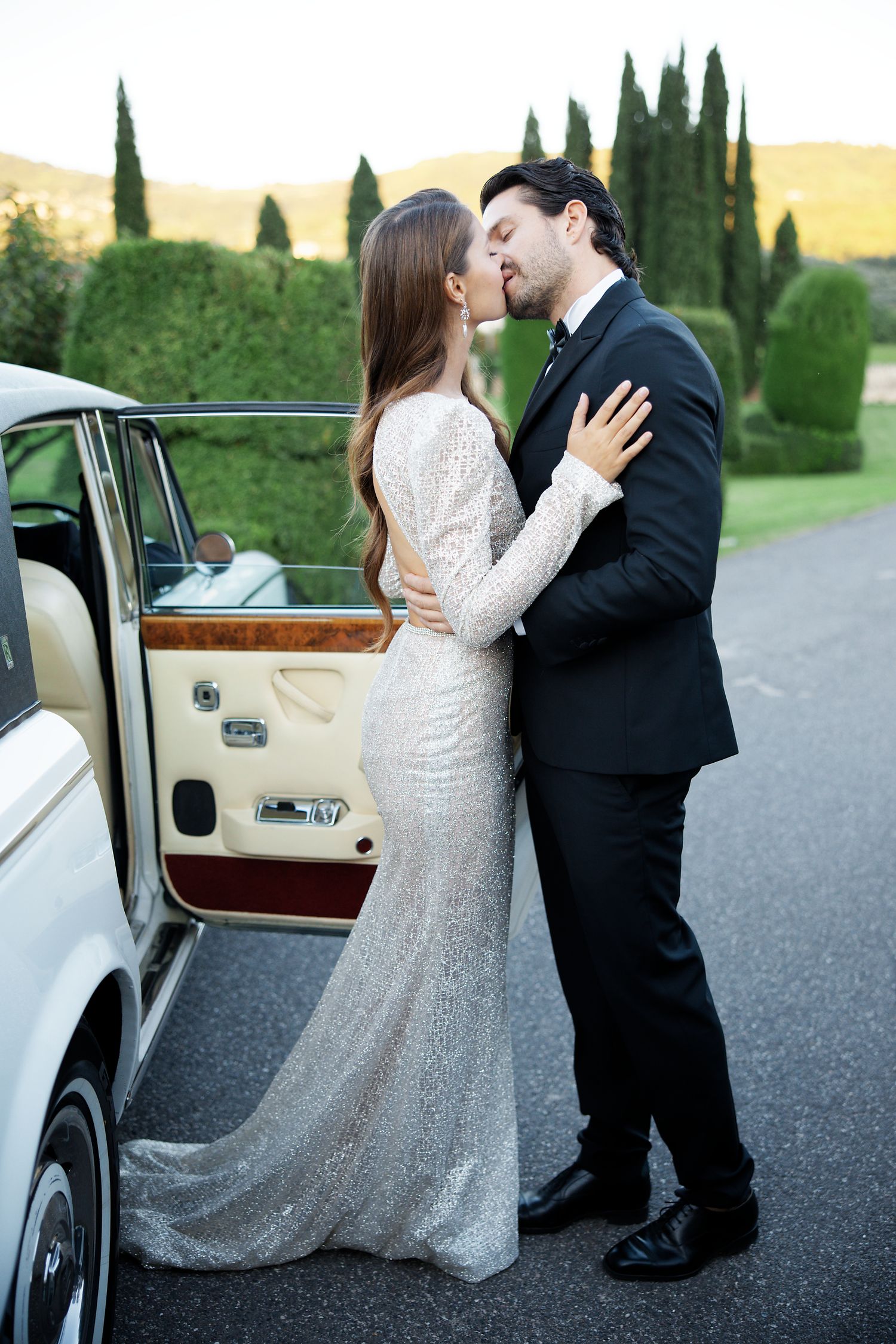 Elegant couple in formal attire embraces near vintage luxury car at sunset.