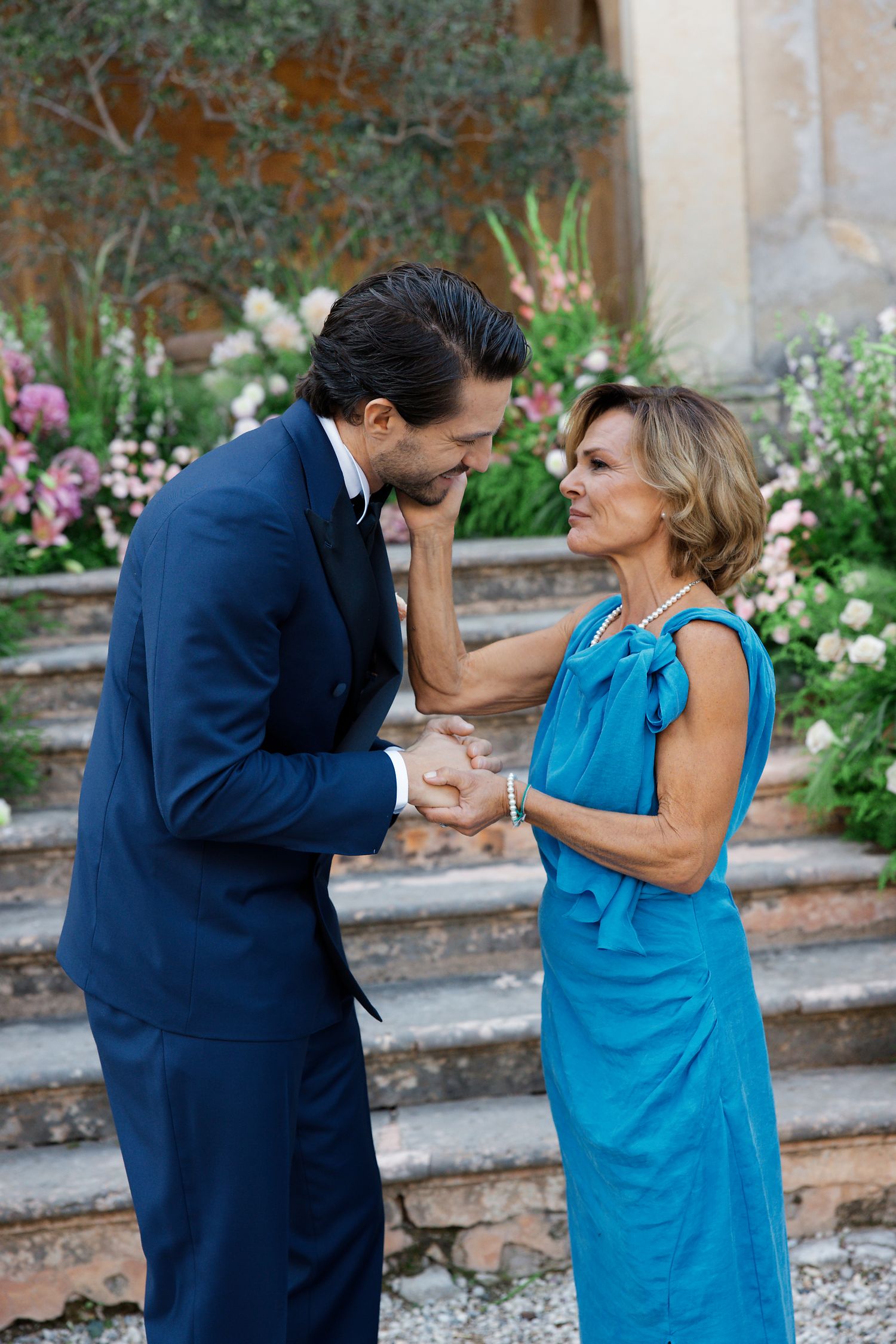 A sweet moment between two people in formal attire standing on garden steps, with the person in blue receiving a hand kiss.