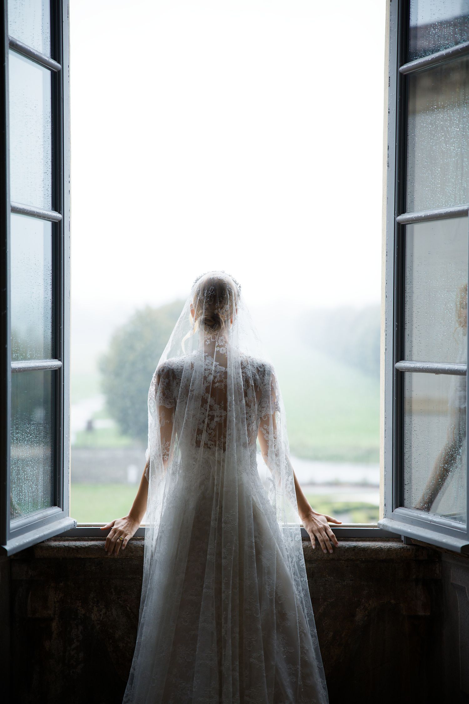 A silhouetted figure in a wedding gown and veil stands at a window overlooking a misty landscape.