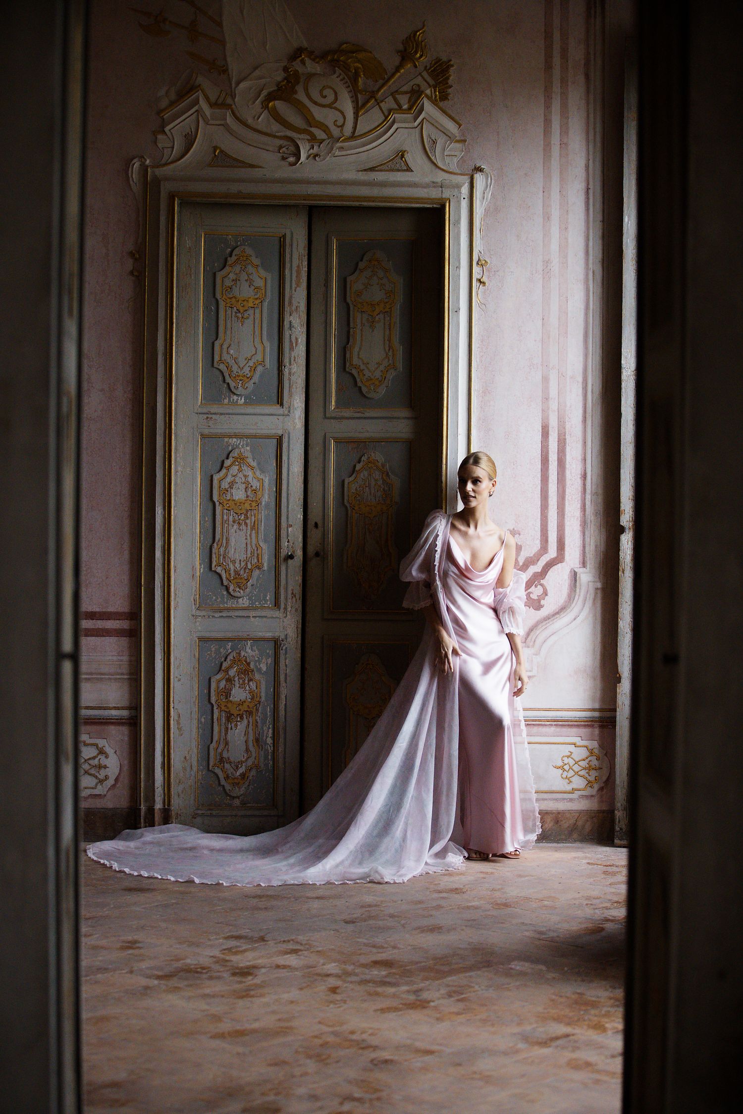 Romantic portrait in a vintage room with ornate doors showing a flowing pink gown with long train.
