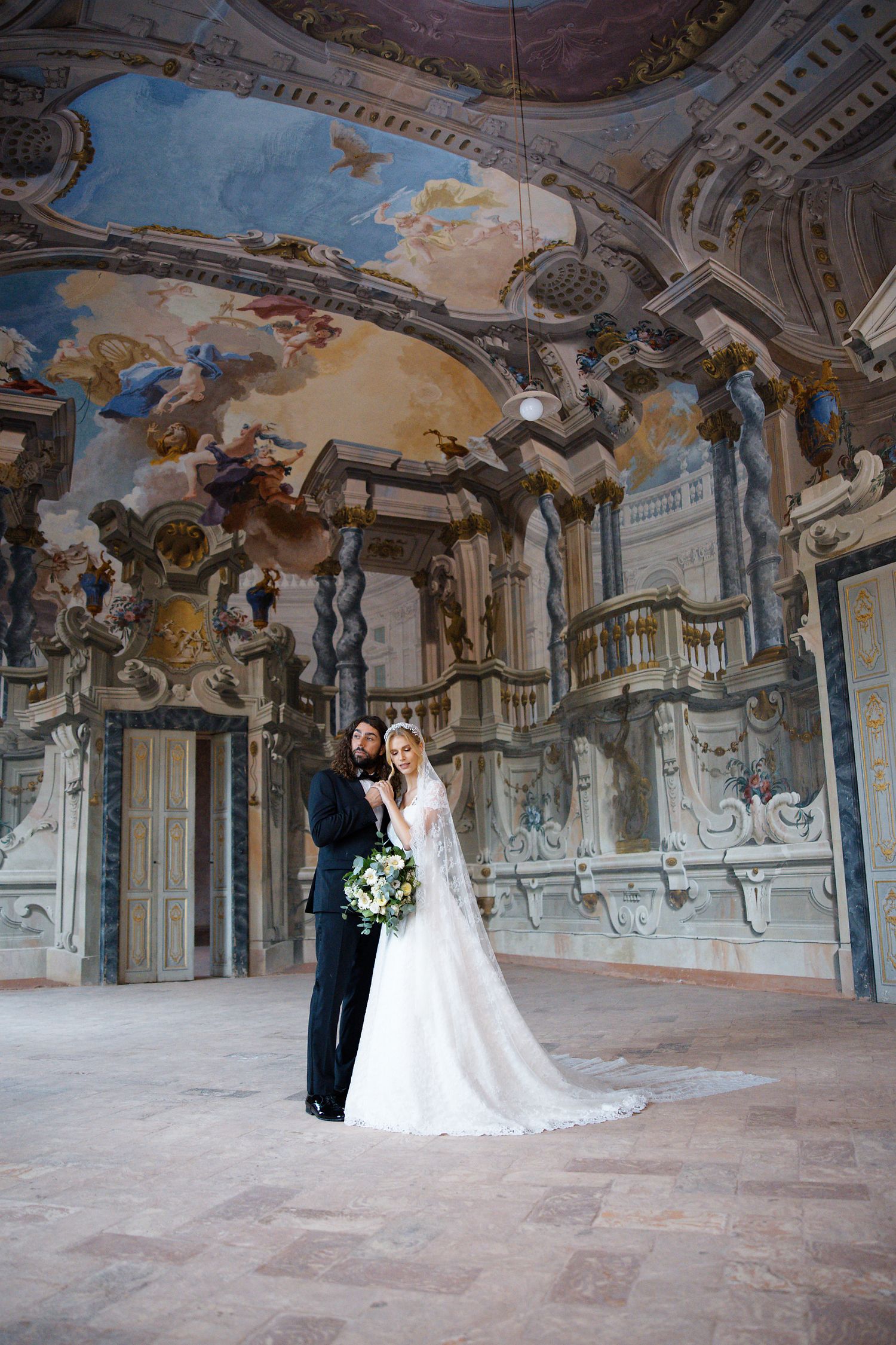 Wedding couple stands beneath baroque ceiling frescoes in grand historic palace.