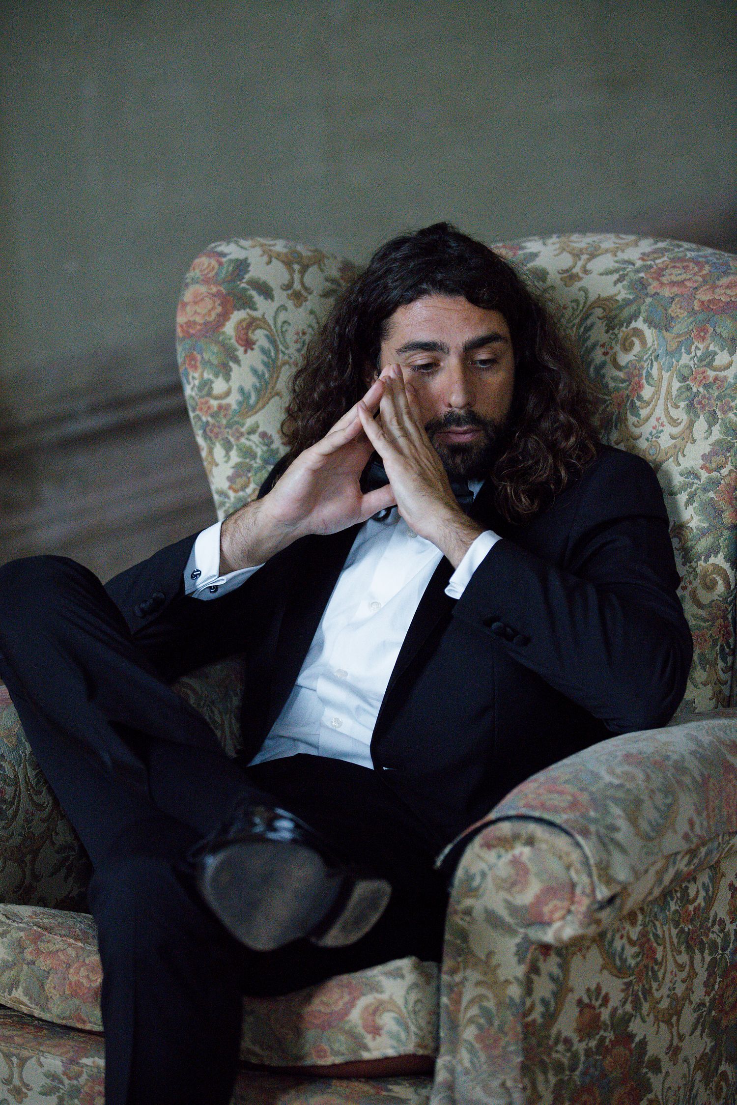 Man in black suit reclines thoughtfully in vintage floral armchair.