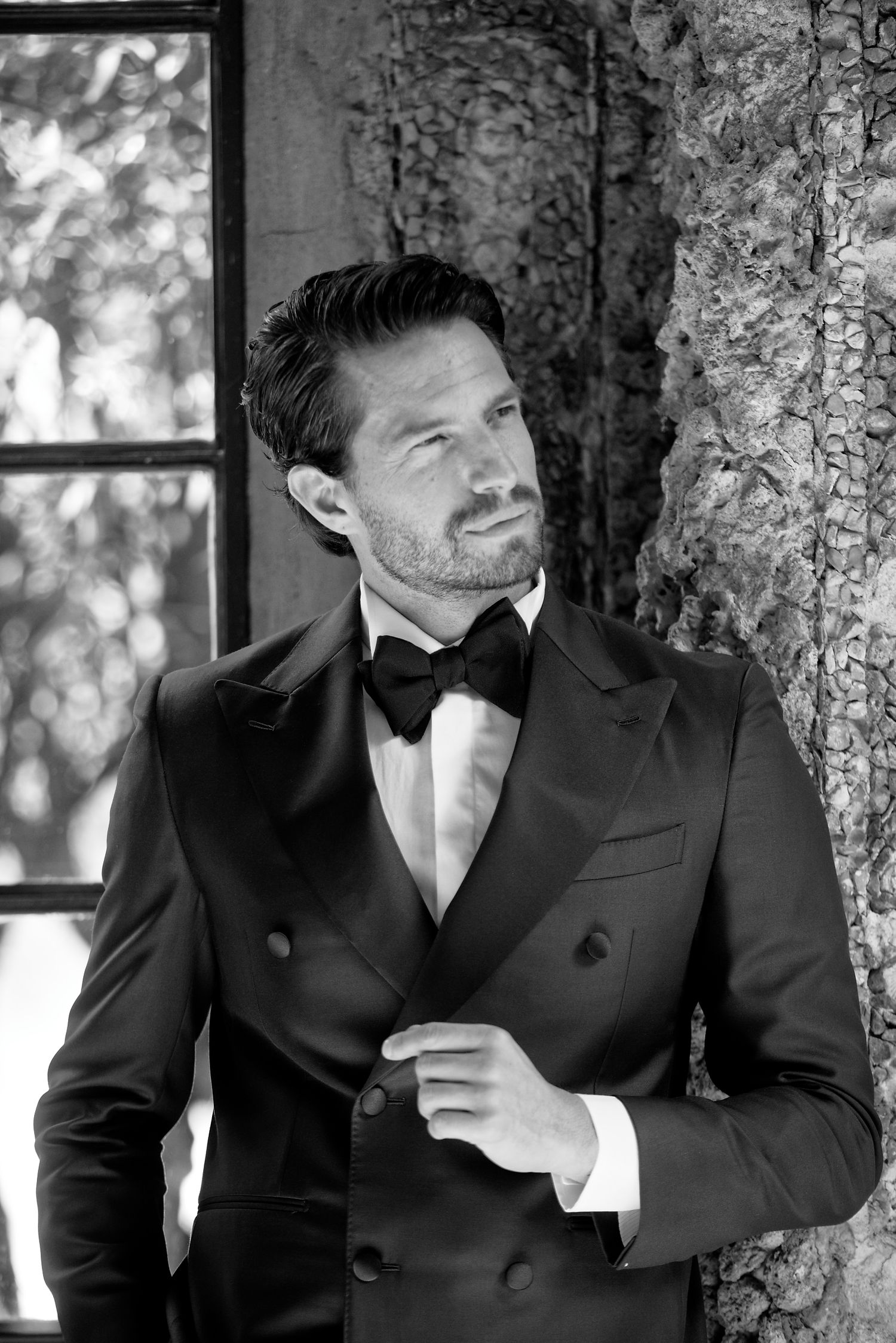 Black and white portrait of someone wearing a black double-breasted tuxedo with bow tie in an elegant setting.