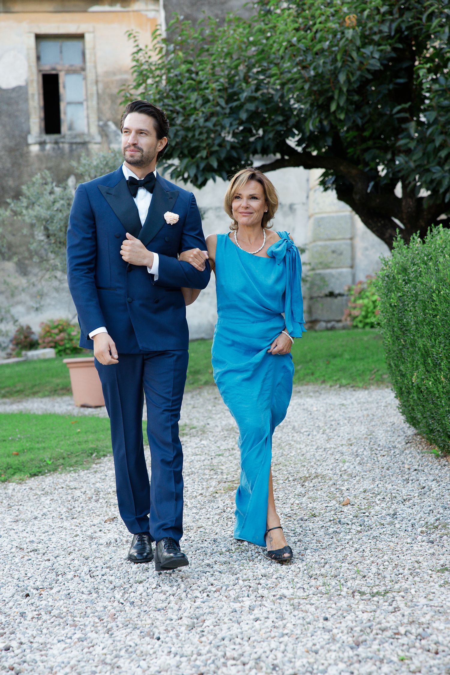 Formal couple walking arm in arm on gravel path, wearing navy suit and turquoise evening dress in garden setting.