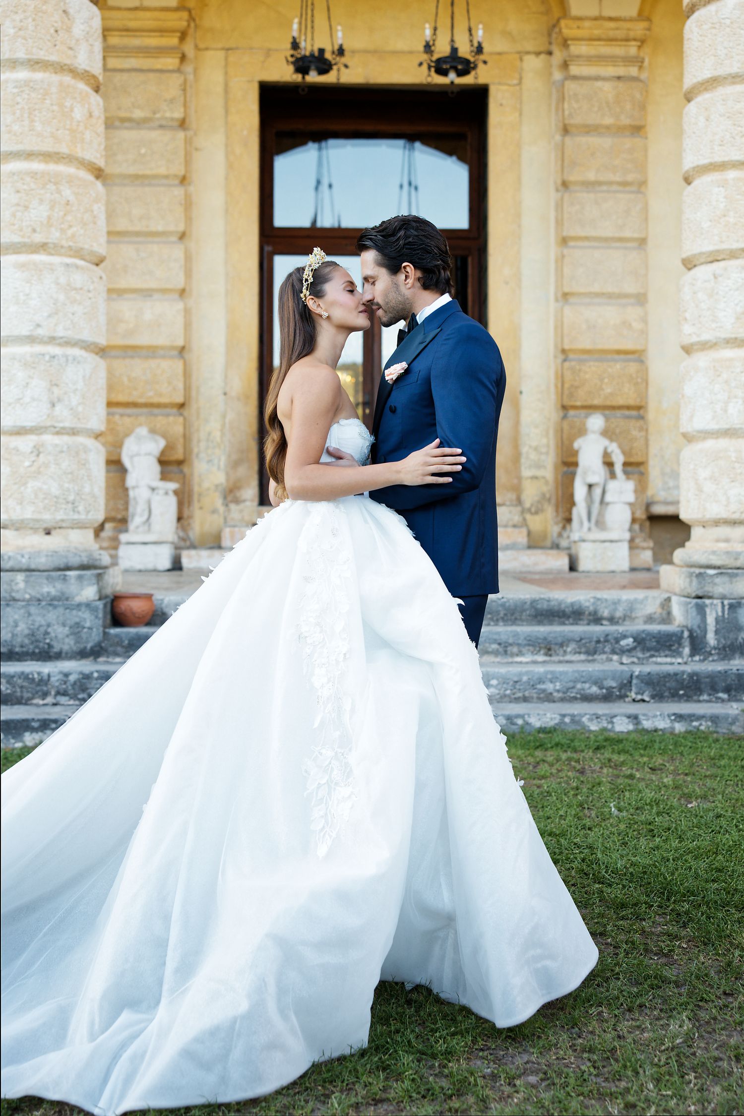 Newlyweds share romantic moment in front of elegant stone mansion with flowing white wedding gown.