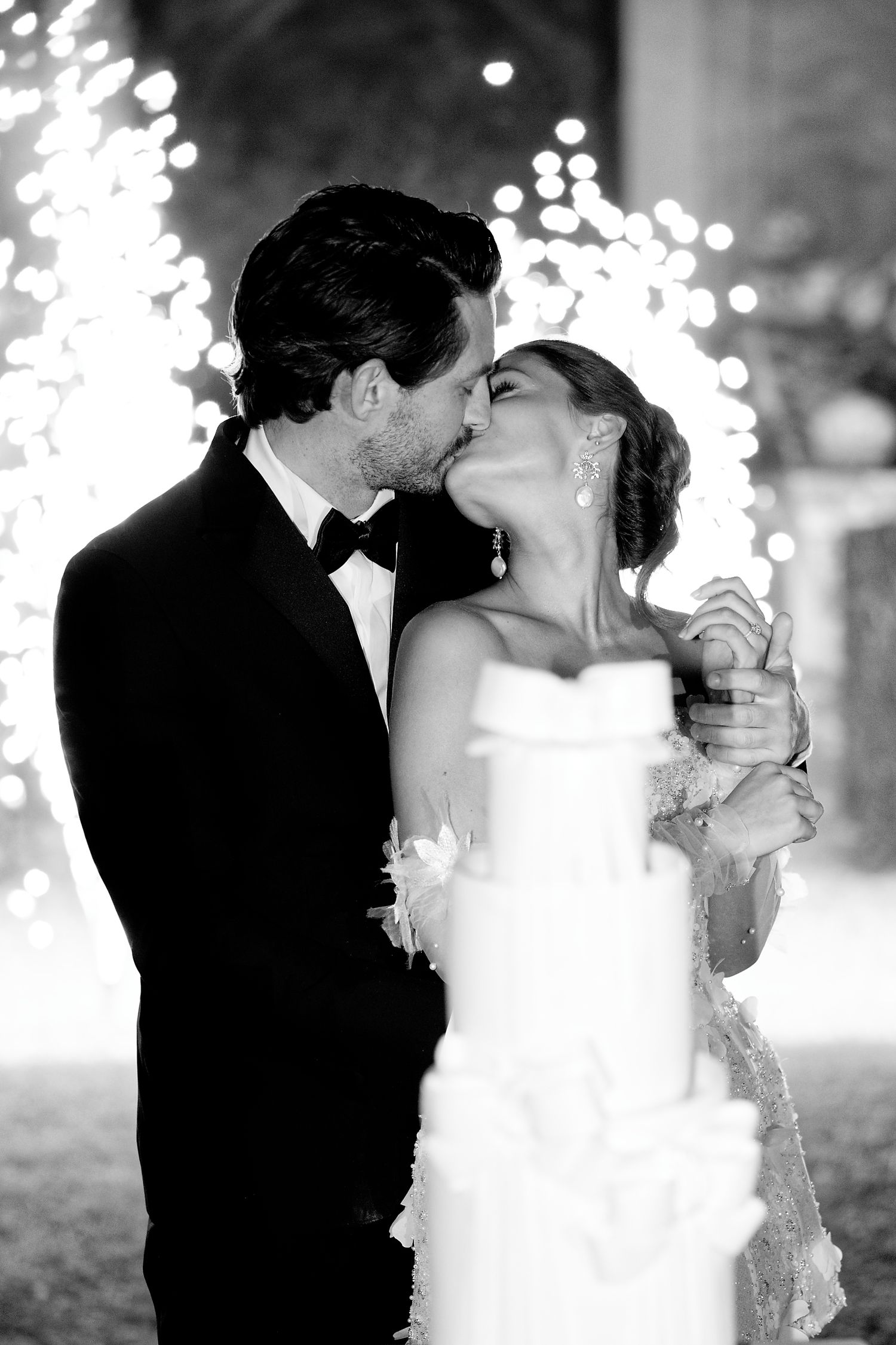 Black and white wedding photo of couple sharing intimate kiss with sparkling lights in background.
