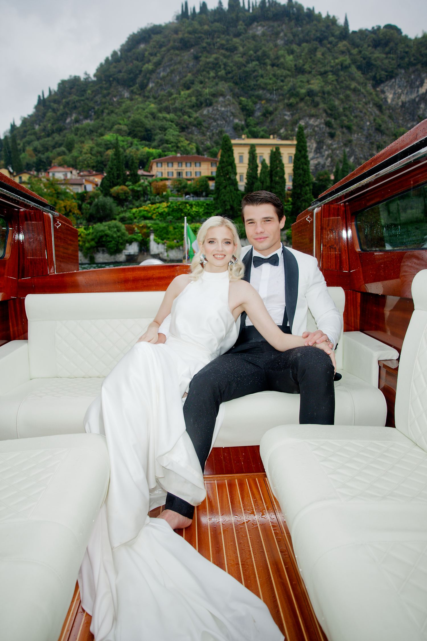 Wedding couple relaxes in white boat seating with mountain villas of Lake Como visible behind them.
