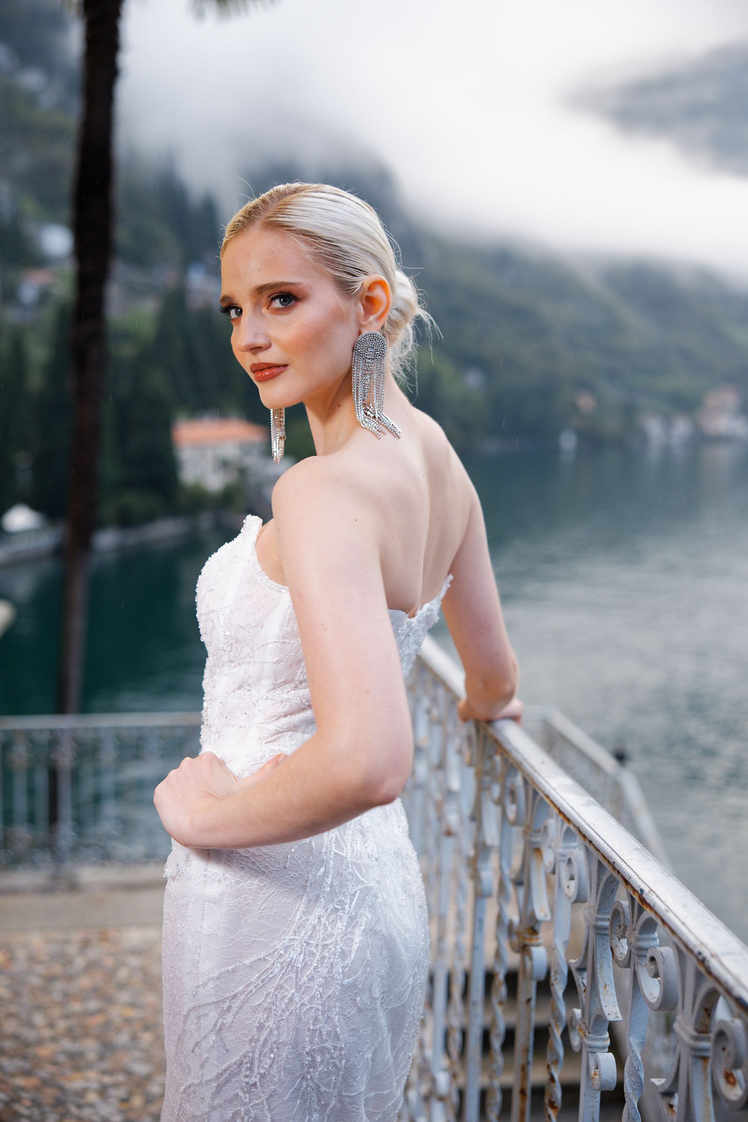 Bride in strapless white gown poses on lakeside terrace with misty mountain views.