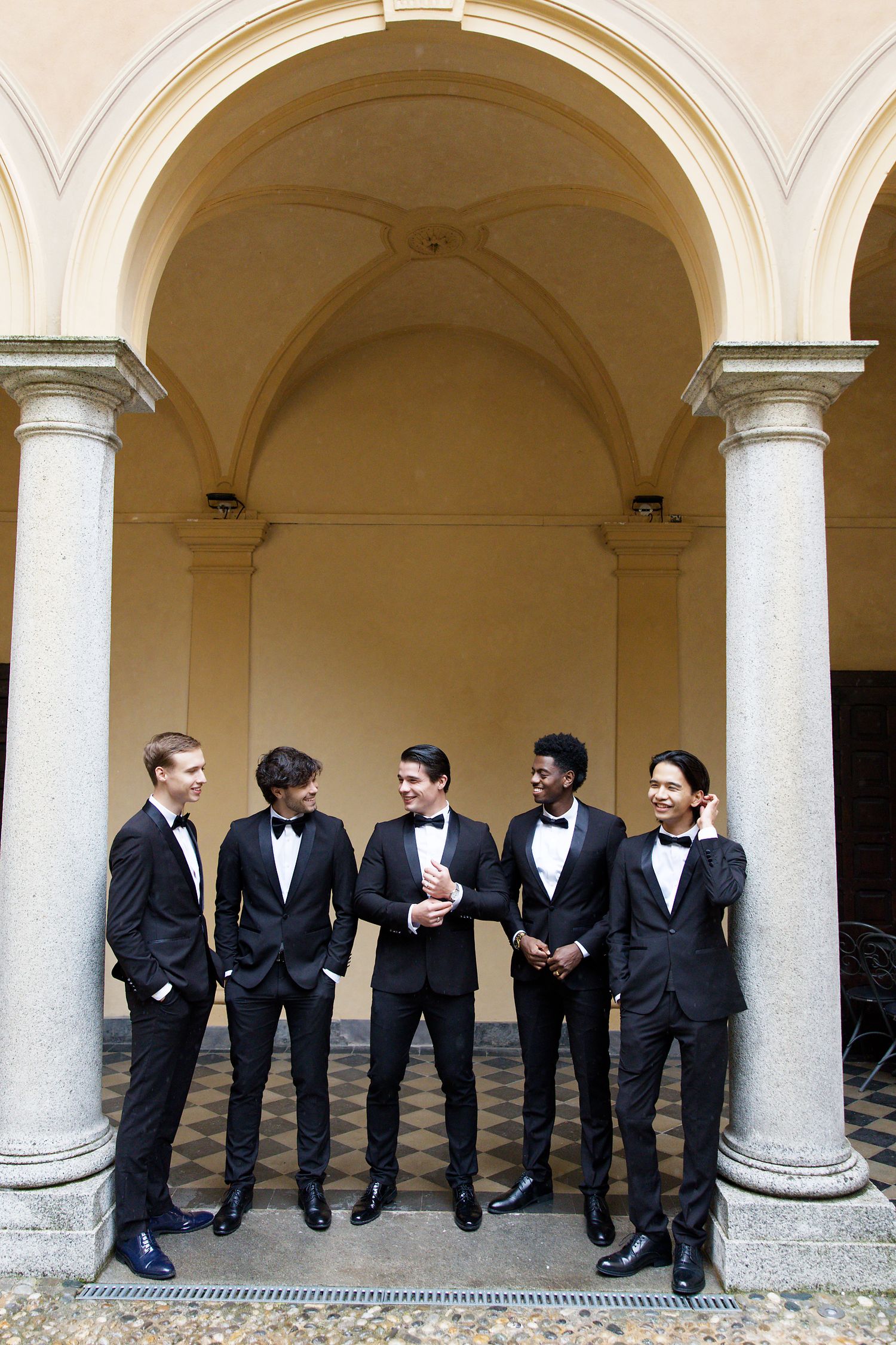 Five groomsmen in black tuxedos stand together under an arched colonnade.