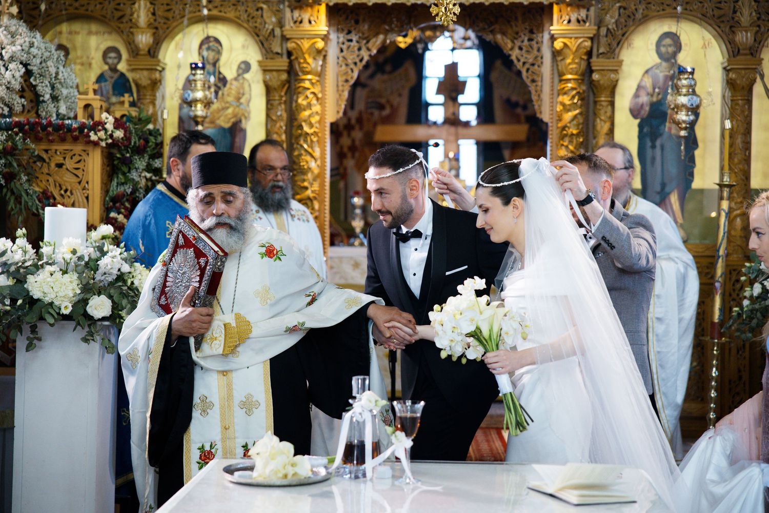 Religious wedding ceremony taking place in an ornate Orthodox church with gold icons and white floral decorations.