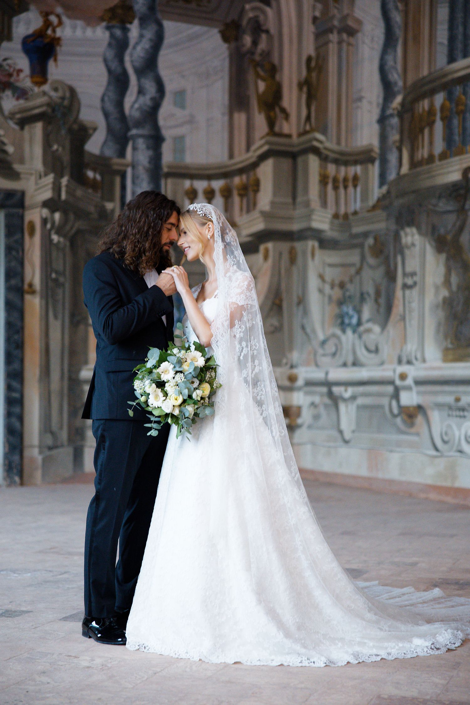 A romantic wedding photo of newlyweds embracing in an elegant baroque-style palace interior.