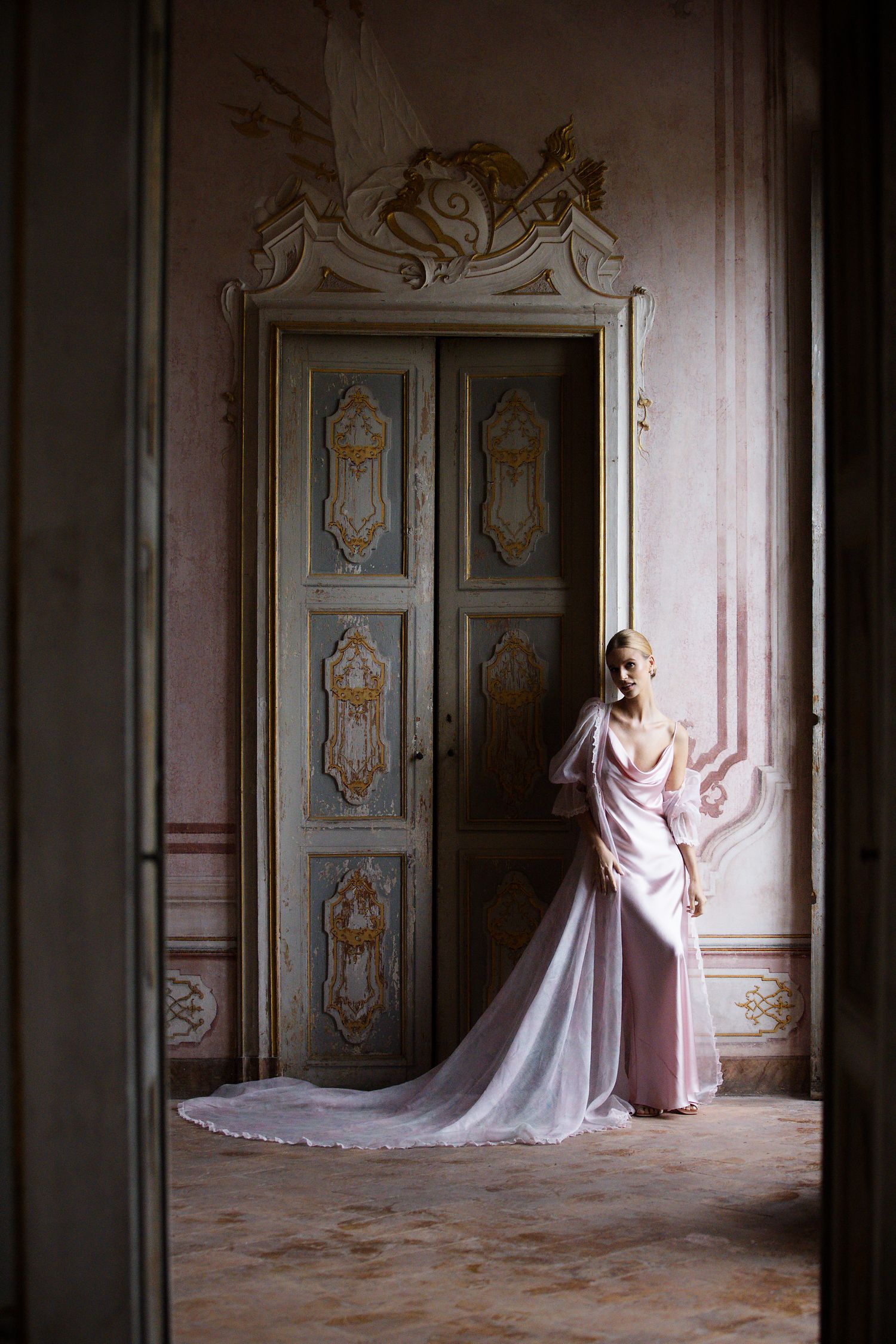 A dramatic full-length shot of a pink gown with train against an ornately carved vintage door.
