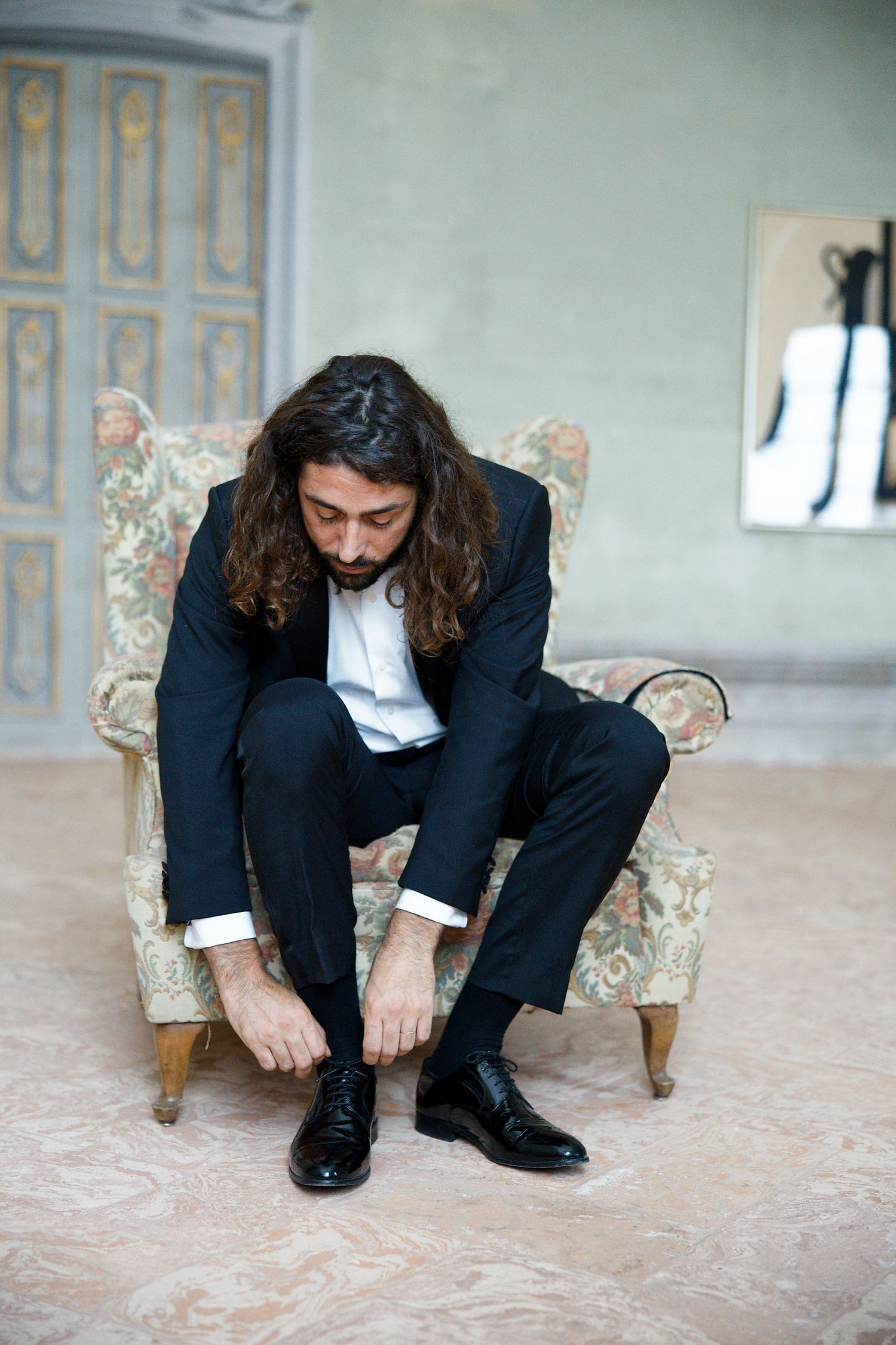 Person in navy suit sitting on vintage floral armchair while tying leather dress shoes.