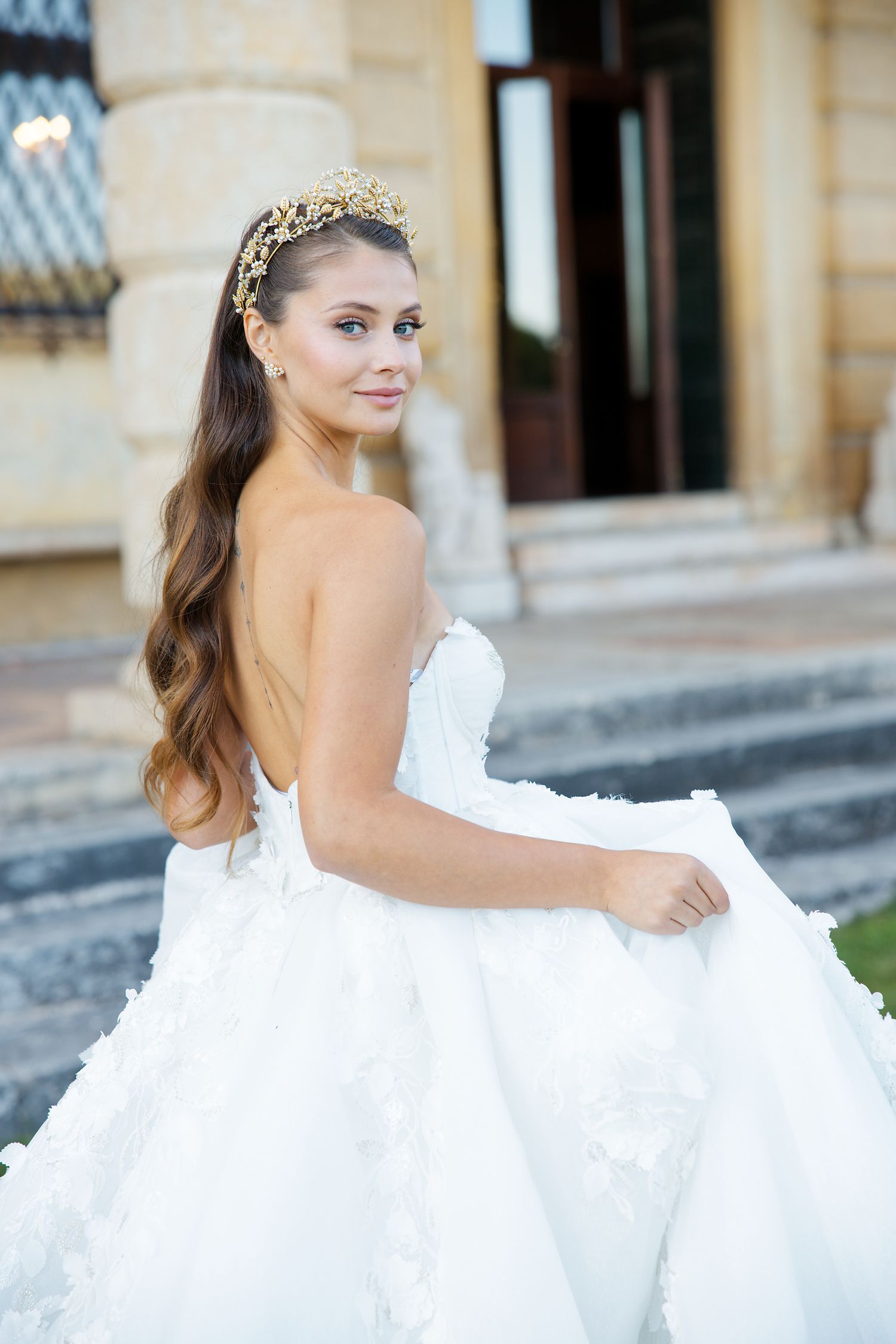 A bride in a flowing white strapless gown and crystal tiara poses gracefully on stone steps.