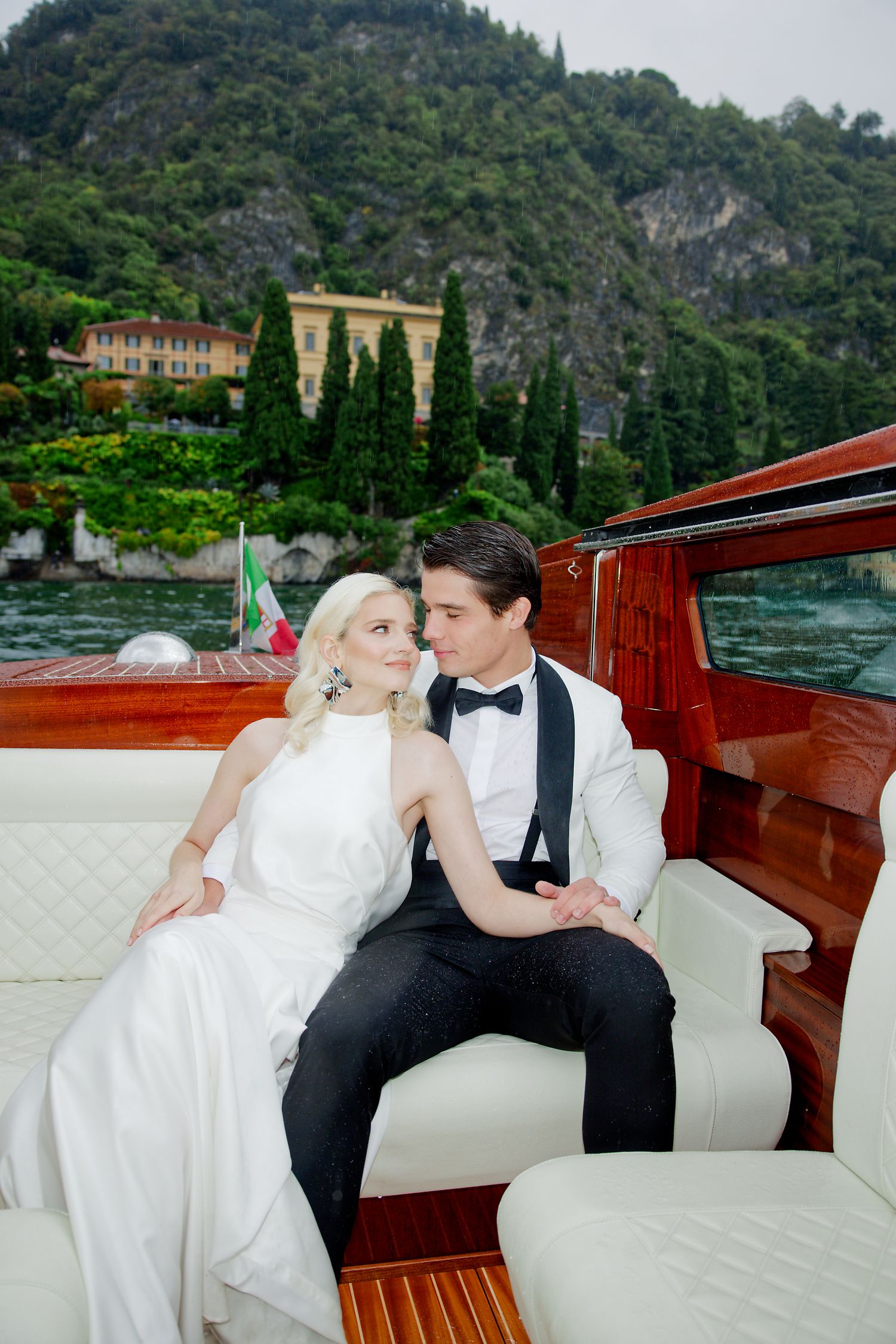 A couple in wedding attire share an intimate moment aboard a wooden boat on a scenic lake in Italy.