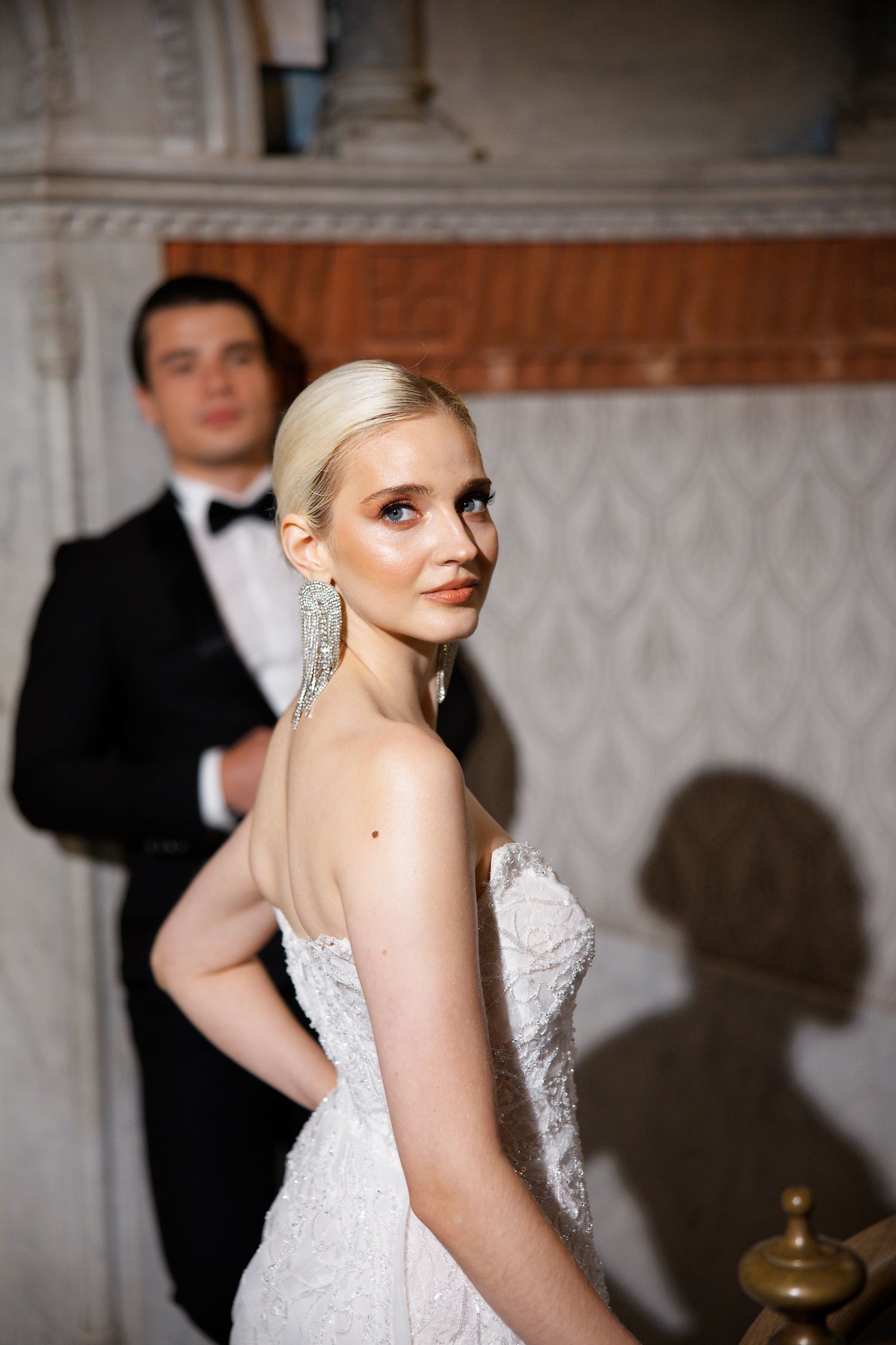 A wedding couple poses in formal attire against an elegant interior wall with decorative molding.