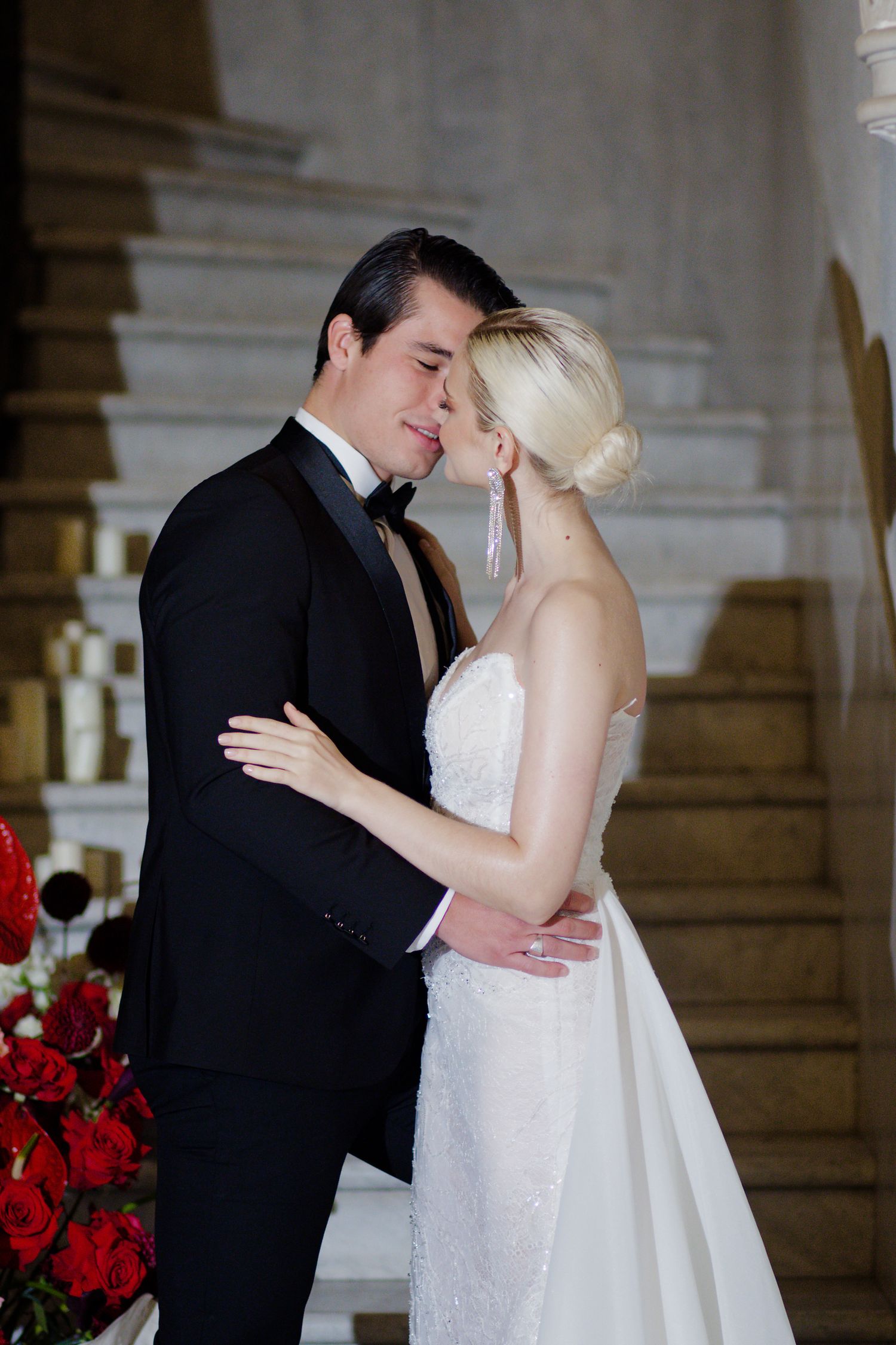 Intimate wedding moment captured by ornate staircase with red floral arrangements.