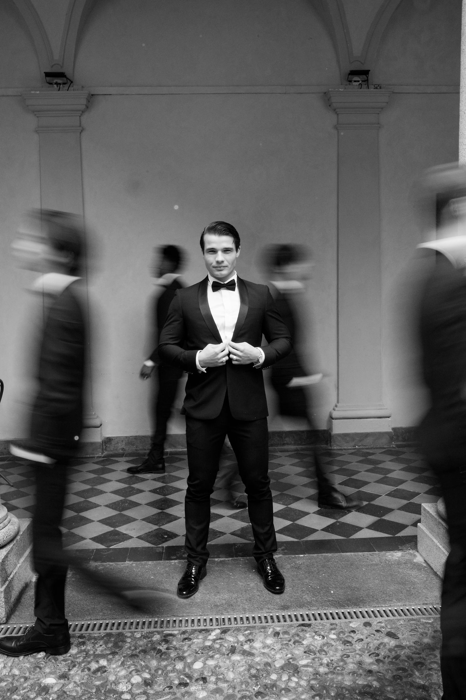 Artistic black and white photo of figure in tuxedo with motion-blurred people walking by in archway.