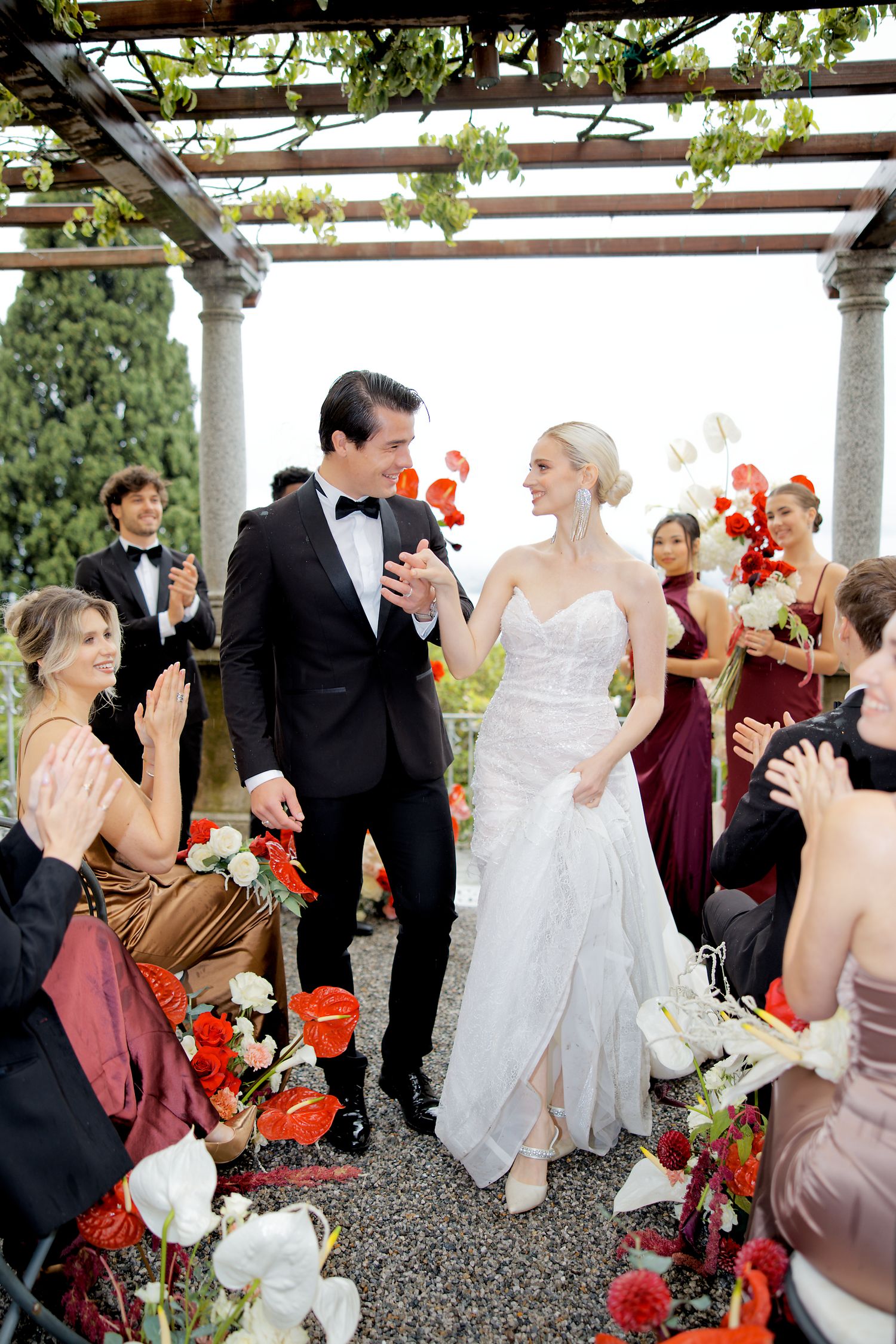 Wedding celebration under a pergola with red and white floral decorations surrounded by guests in formal attire.
