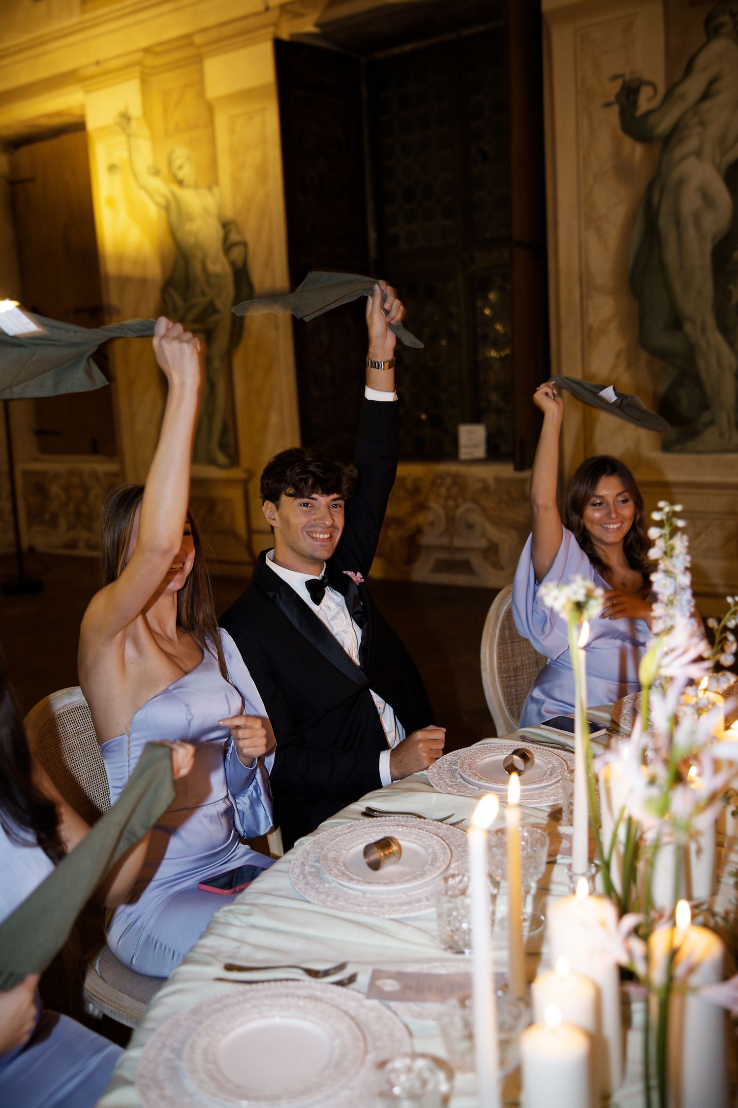 Wedding guests wave napkins joyfully during reception dinner at an elegant candle-lit table.