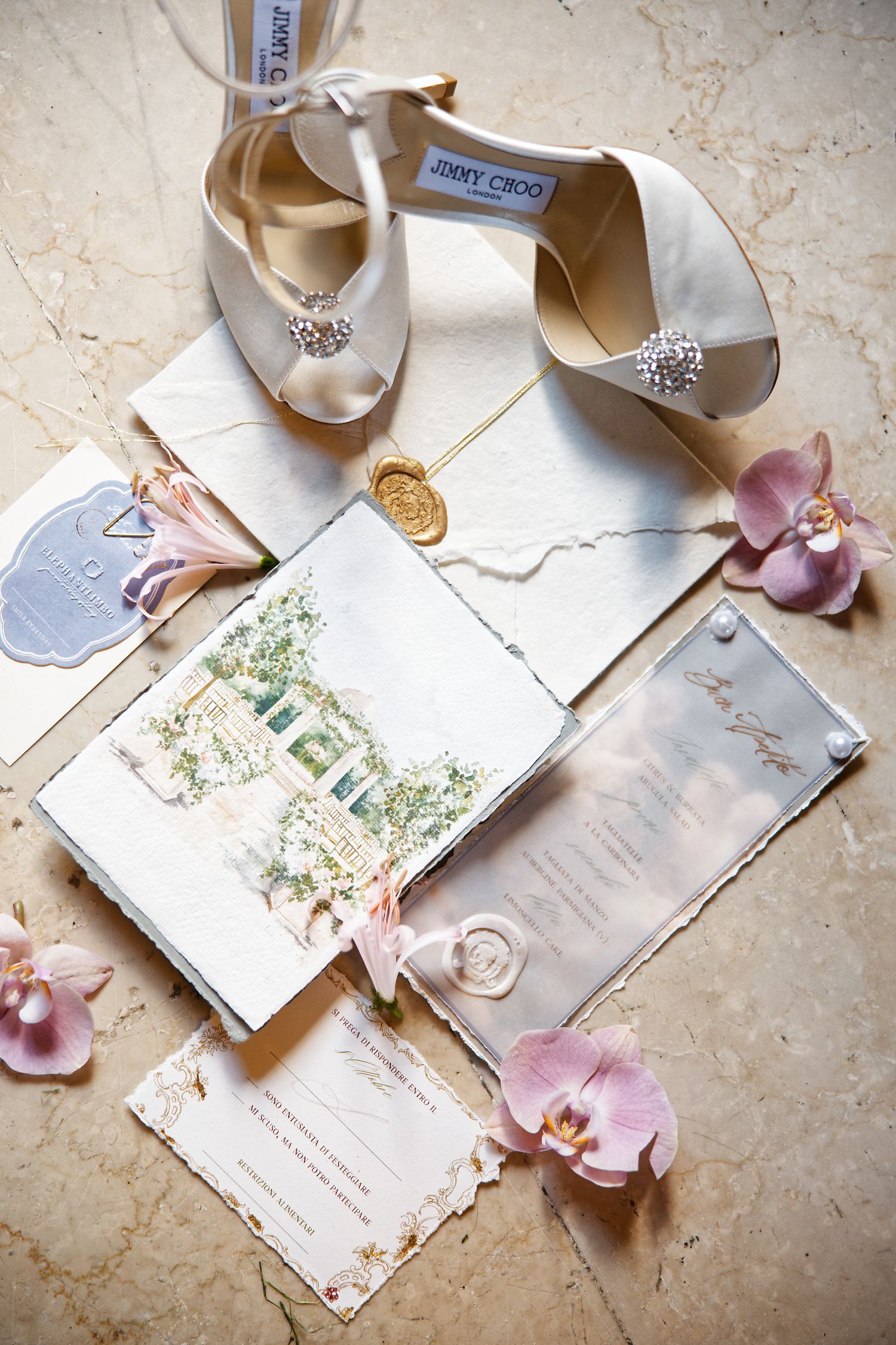 Wedding details featuring Jimmy Choo shoes, invitation cards, and delicate pink orchids on marble surface.