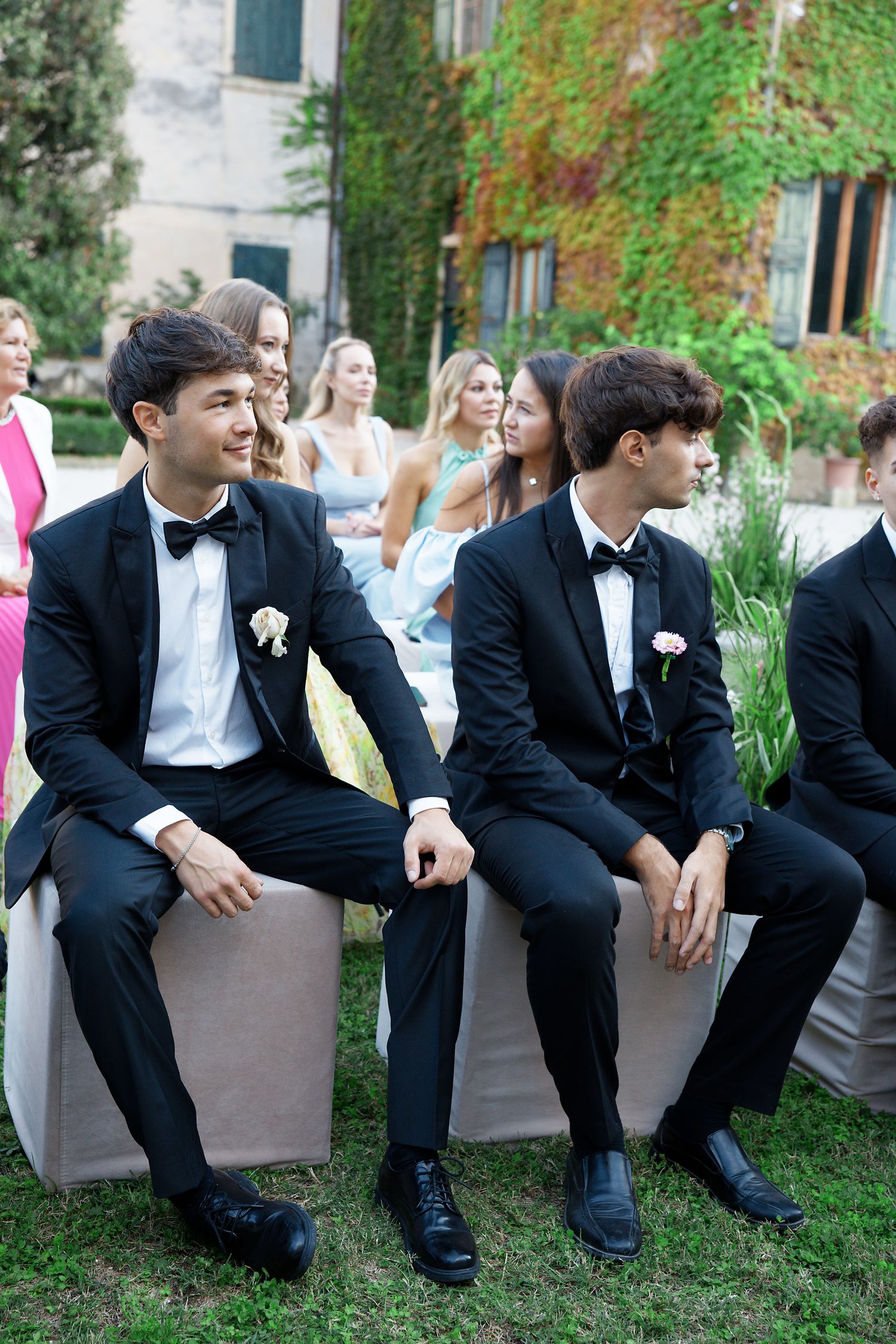 Groomsmen in black tuxedos sit on benches at an outdoor wedding venue with ivy-covered walls.