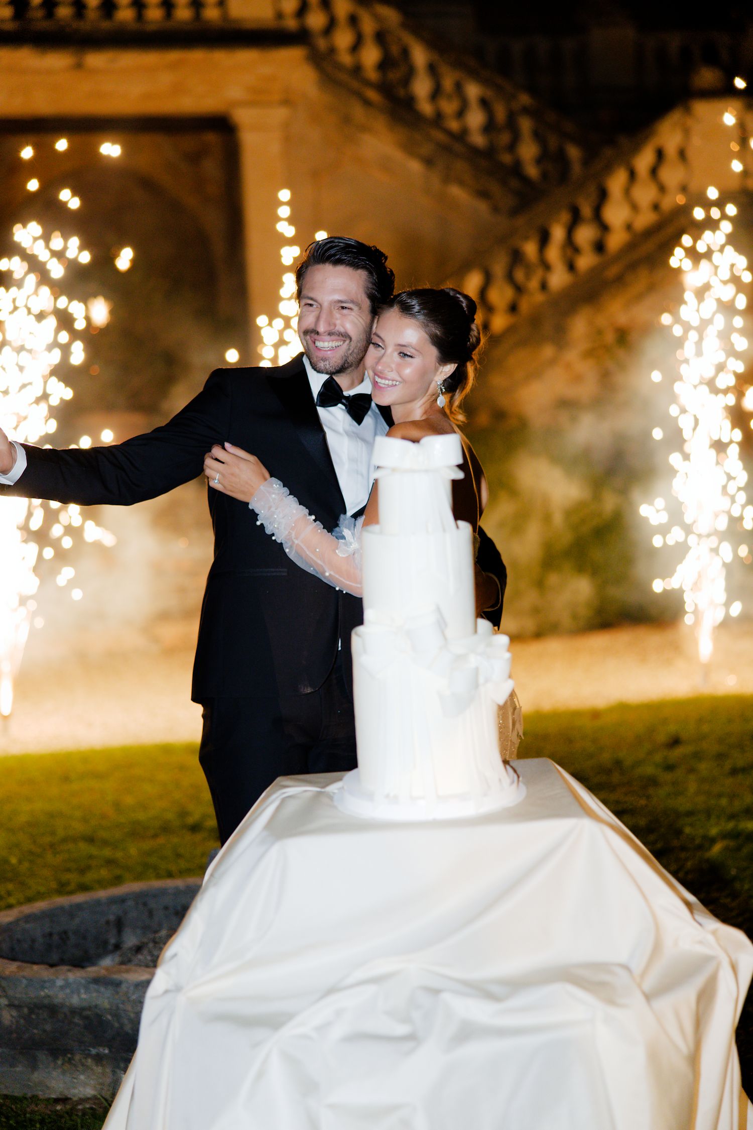 Newlyweds cutting elegant white wedding cake with sparklers glowing in background.
