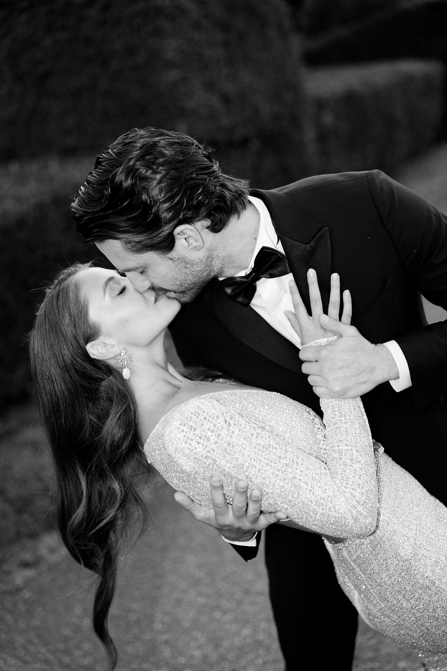 A romantic black and white photo of a couple in formal wear sharing a dip kiss outdoors.