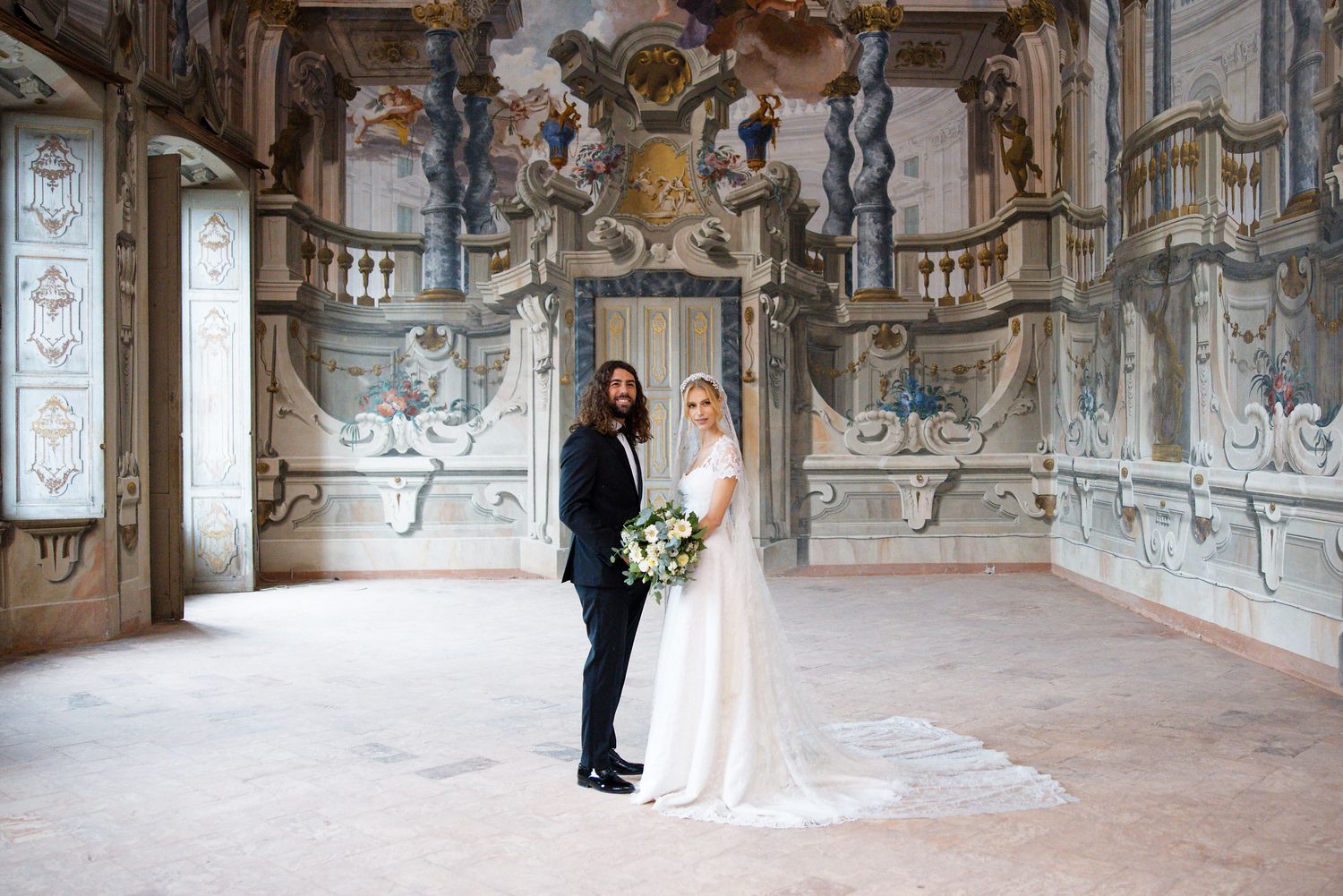 Two people celebrate their wedding in an ornate baroque hall with frescoed walls and classical architectural details.