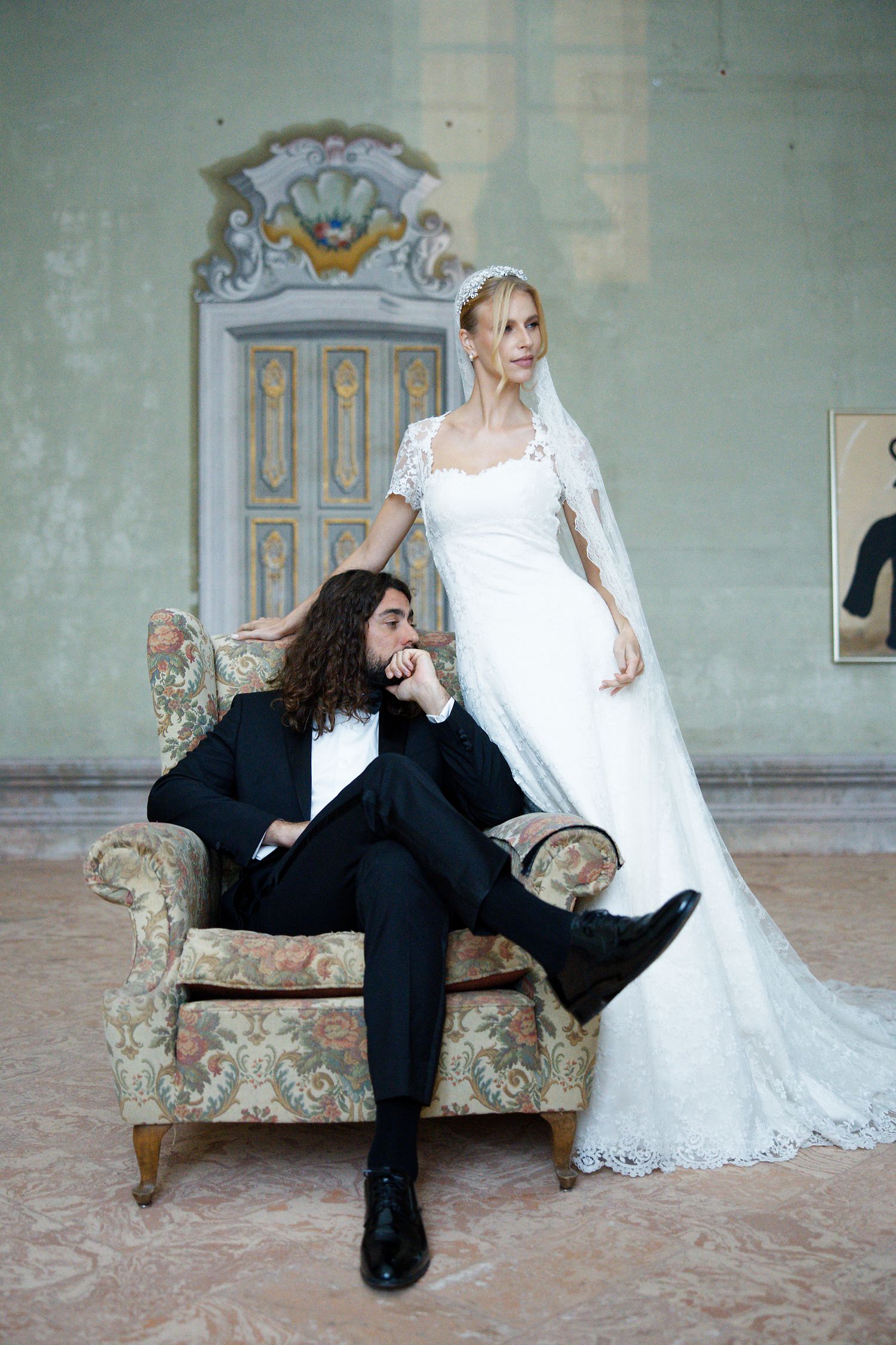 Wedding portrait featuring an elegant white lace gown and figure in black suit lounging on vintage floral chair against mint walls.