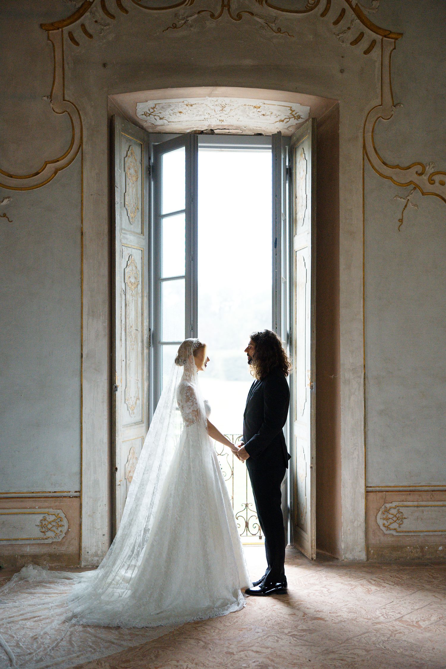 Silhouette of couple in elegant wedding attire standing by ornate window in historic setting.