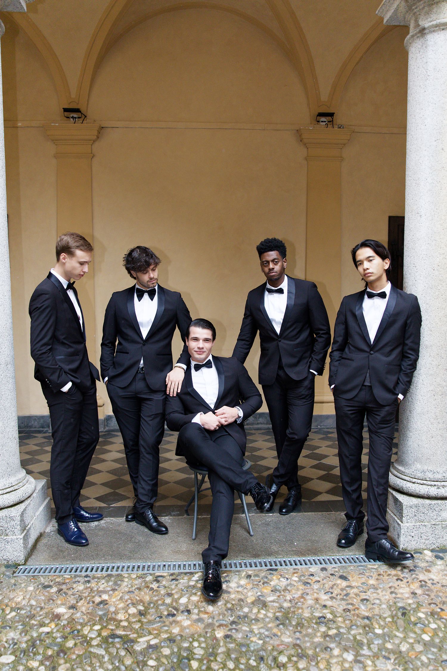 A group of men in black tuxedos and bow ties pose together in an elegant arched hallway with stone columns.