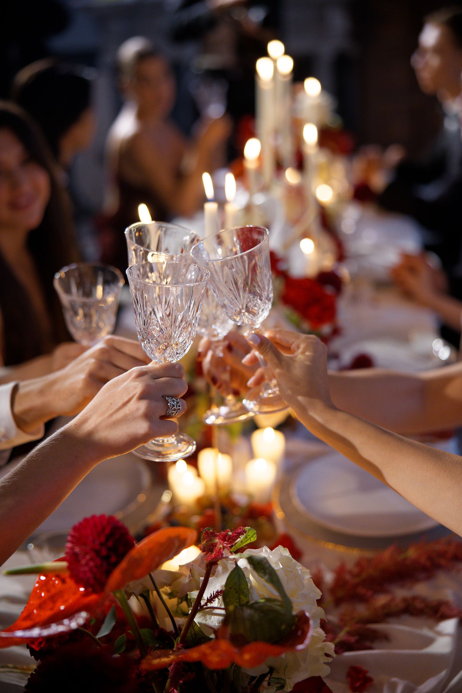 Crystal glasses clink together in a celebratory toast over an elegantly set dinner table with candles and floral arrangements.